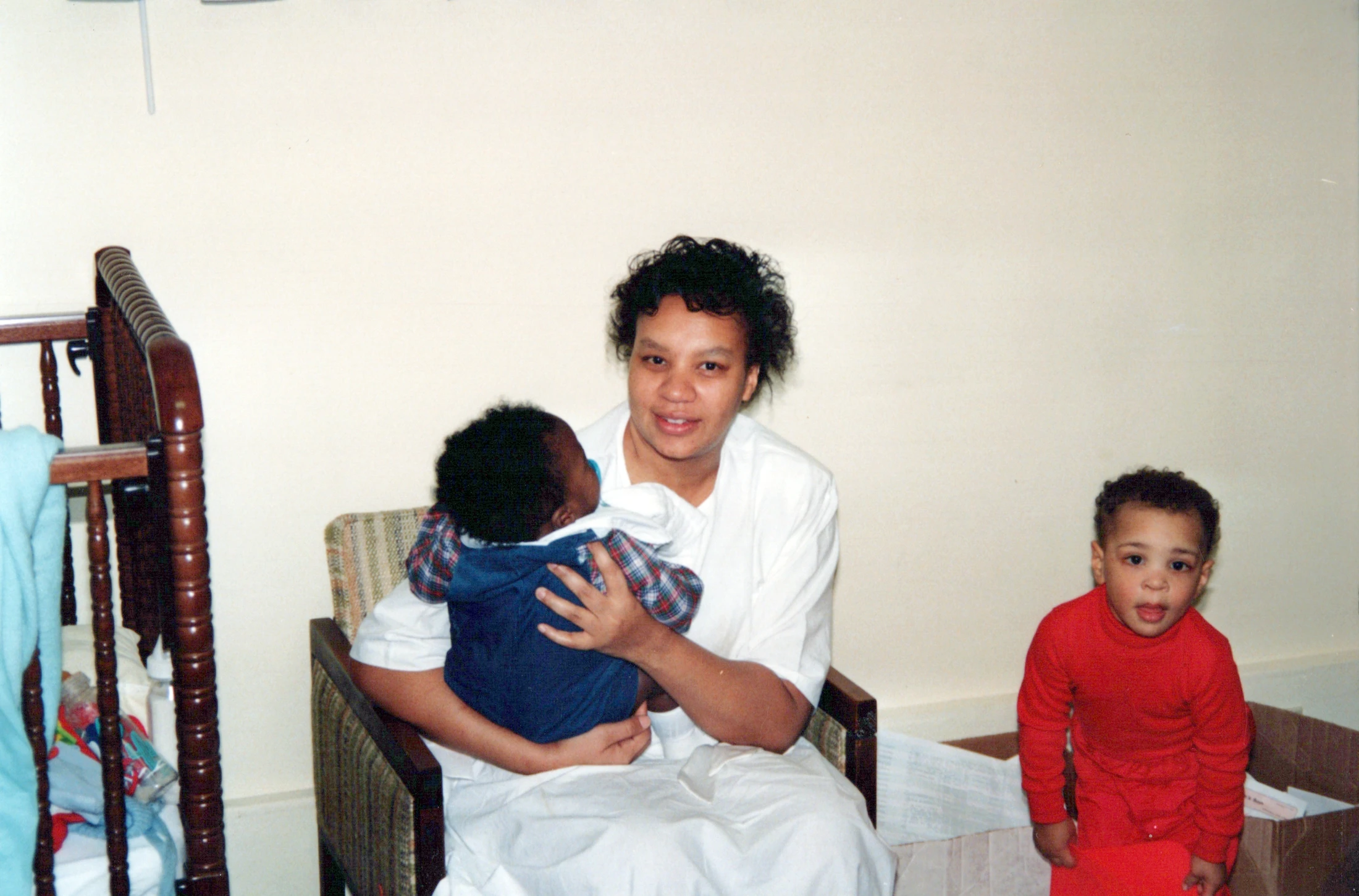 A vintage photograph of an African American woman sitting in a chair holding a baby, with a young toddler in red pajamas standing beside her.