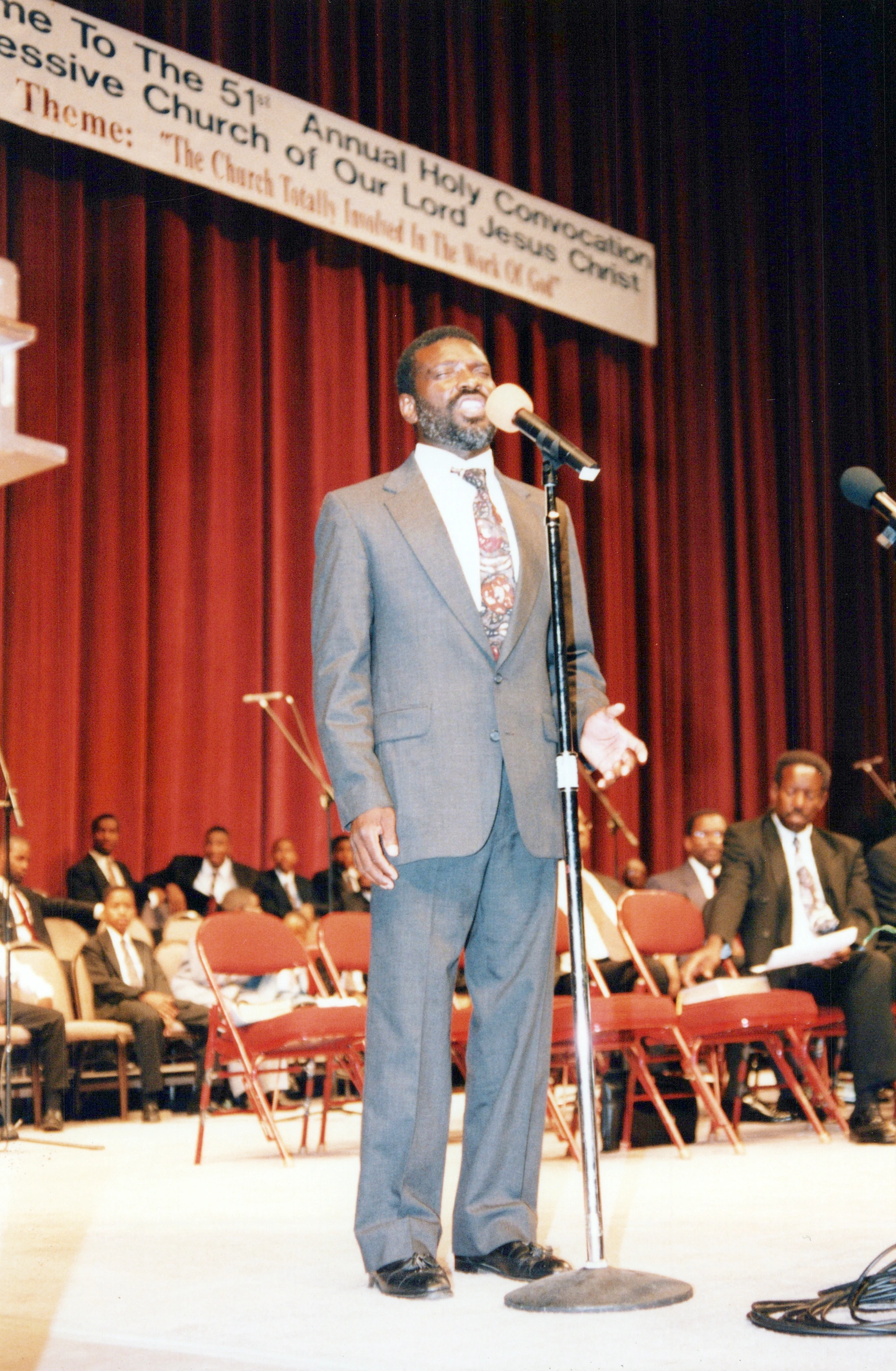A Black man in a grey suit stands on a stage speaking into a microphone during a church convocation. A red curtain and a banner are behind him.