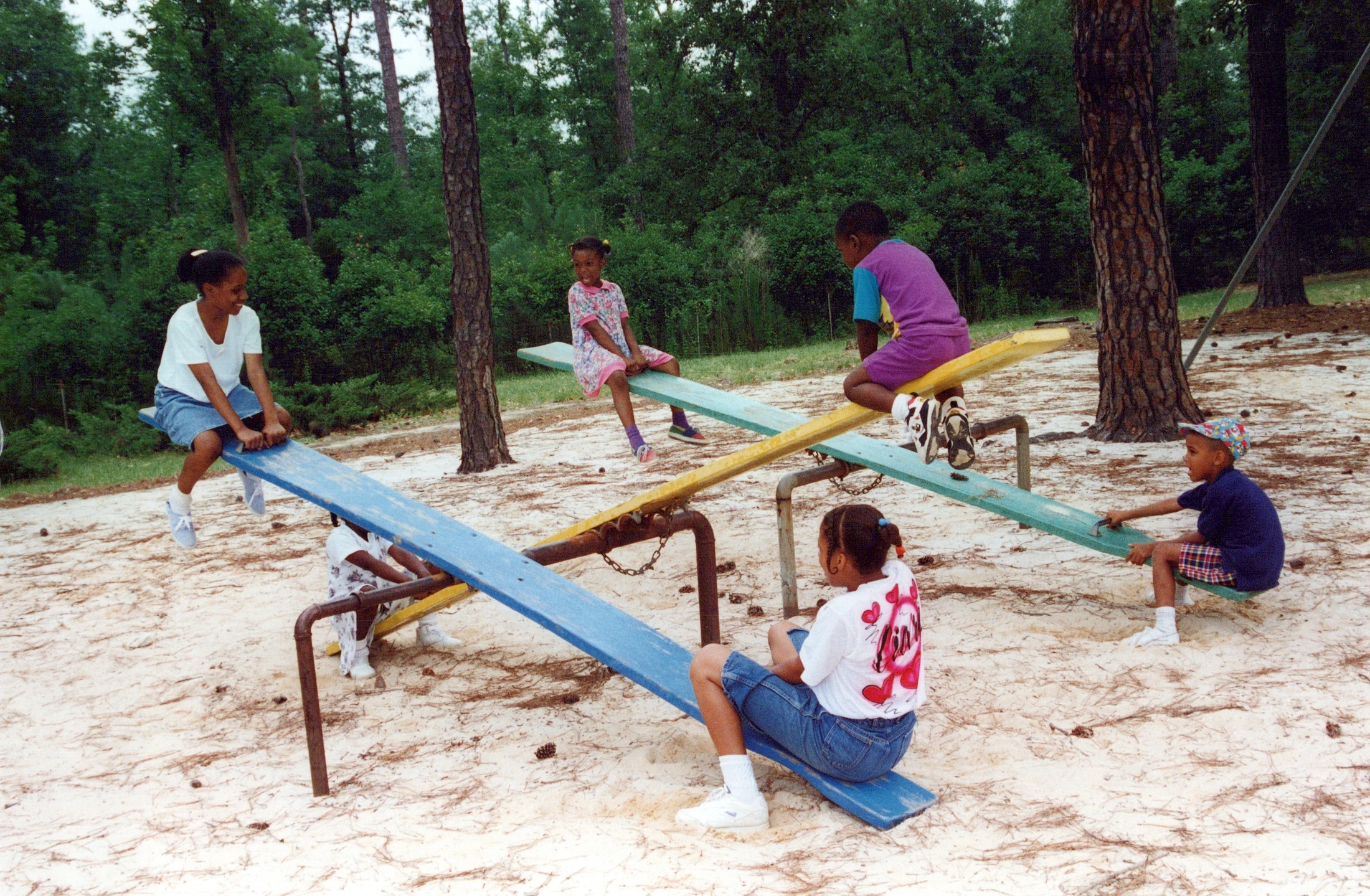 A vintage photograph of six young children playing together on three colorful seesaws in a sandy playground surrounded by pine trees.