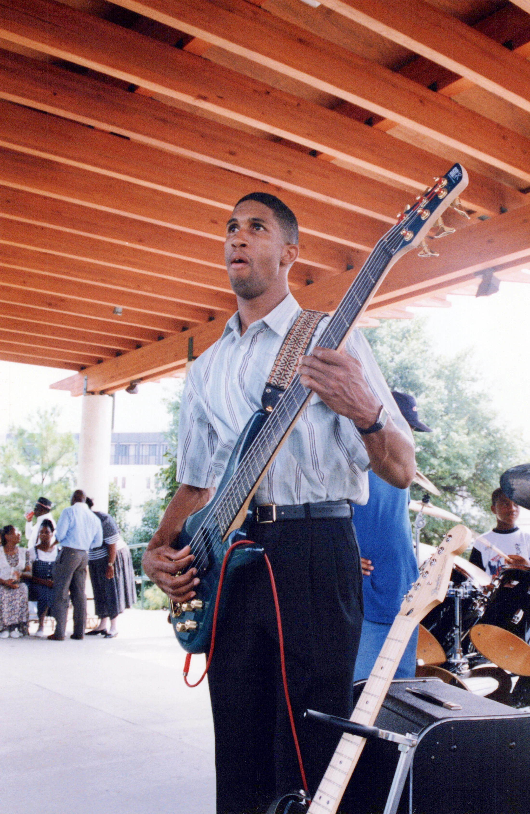 A Black man in a striped shirt plays a bass guitar during an outdoor performance. He stands under a wooden pavilion with a drummer and an audience behind him.