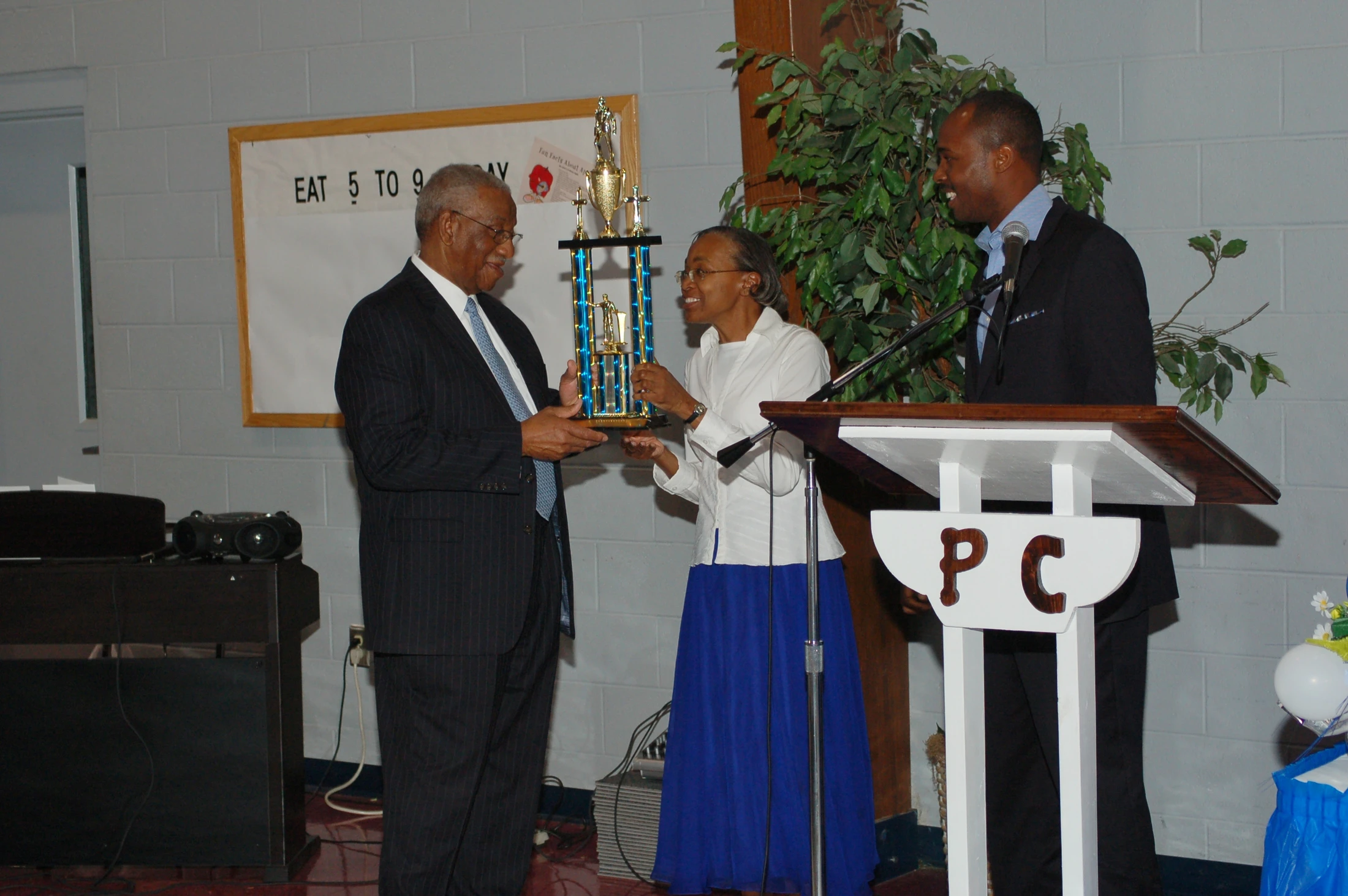 An older man in a suit smiles as he receives a large trophy from a woman, while another man watches from a lectern during an awards ceremony.