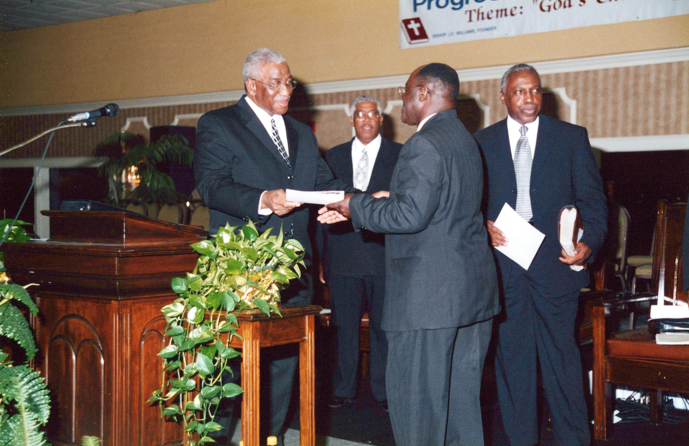An older Black man in a suit hands a certificate to another man over a lectern during a formal church event, as two other men look on.
