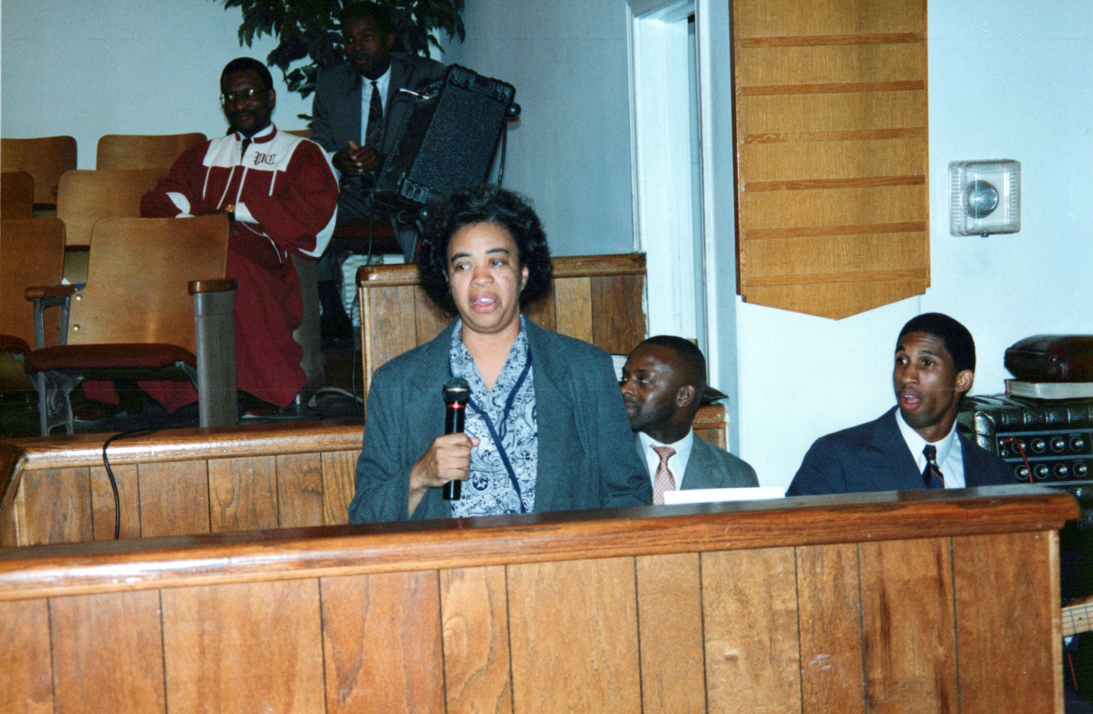 A Black woman in a grey jacket holds a microphone and speaks from a wooden pew. Several men are seated behind her, listening in what appears to be a church.