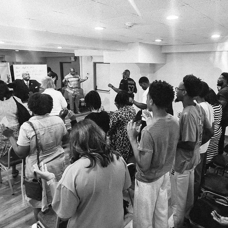 A black and white photograph of a group of people standing in a room during a service, with many raising their hands in prayer and worship.