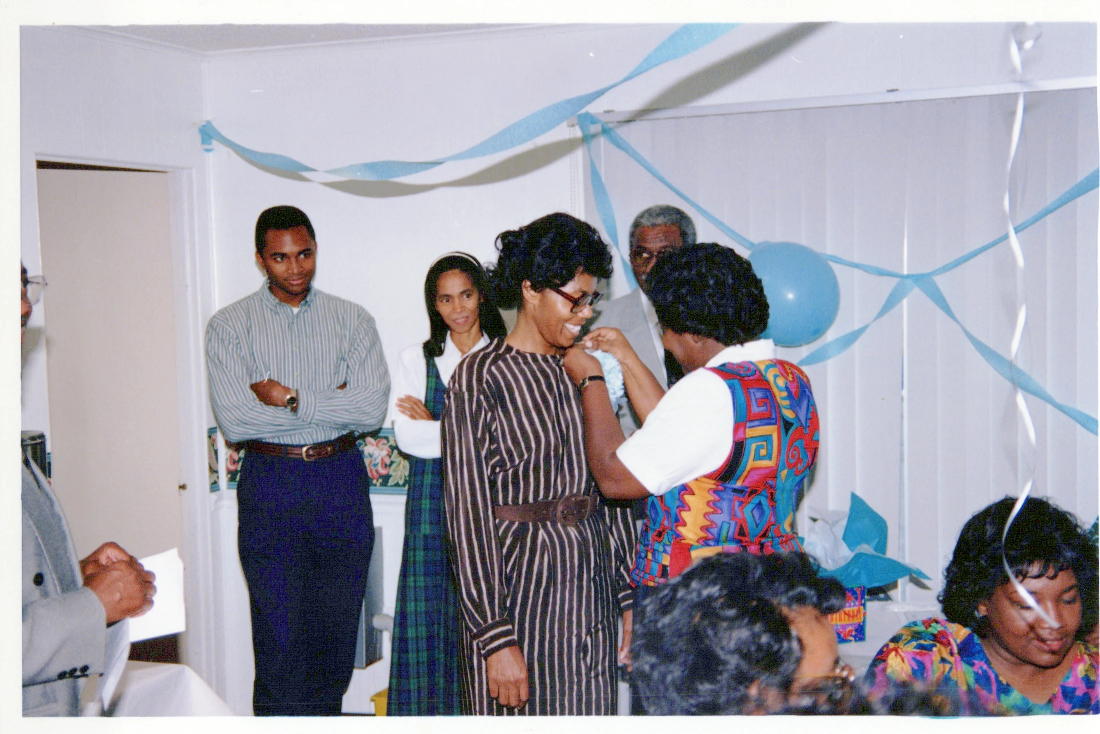 A group of Black people at an indoor party. A woman in a colorful vest pins something on a smiling woman in a striped dress as others look on.