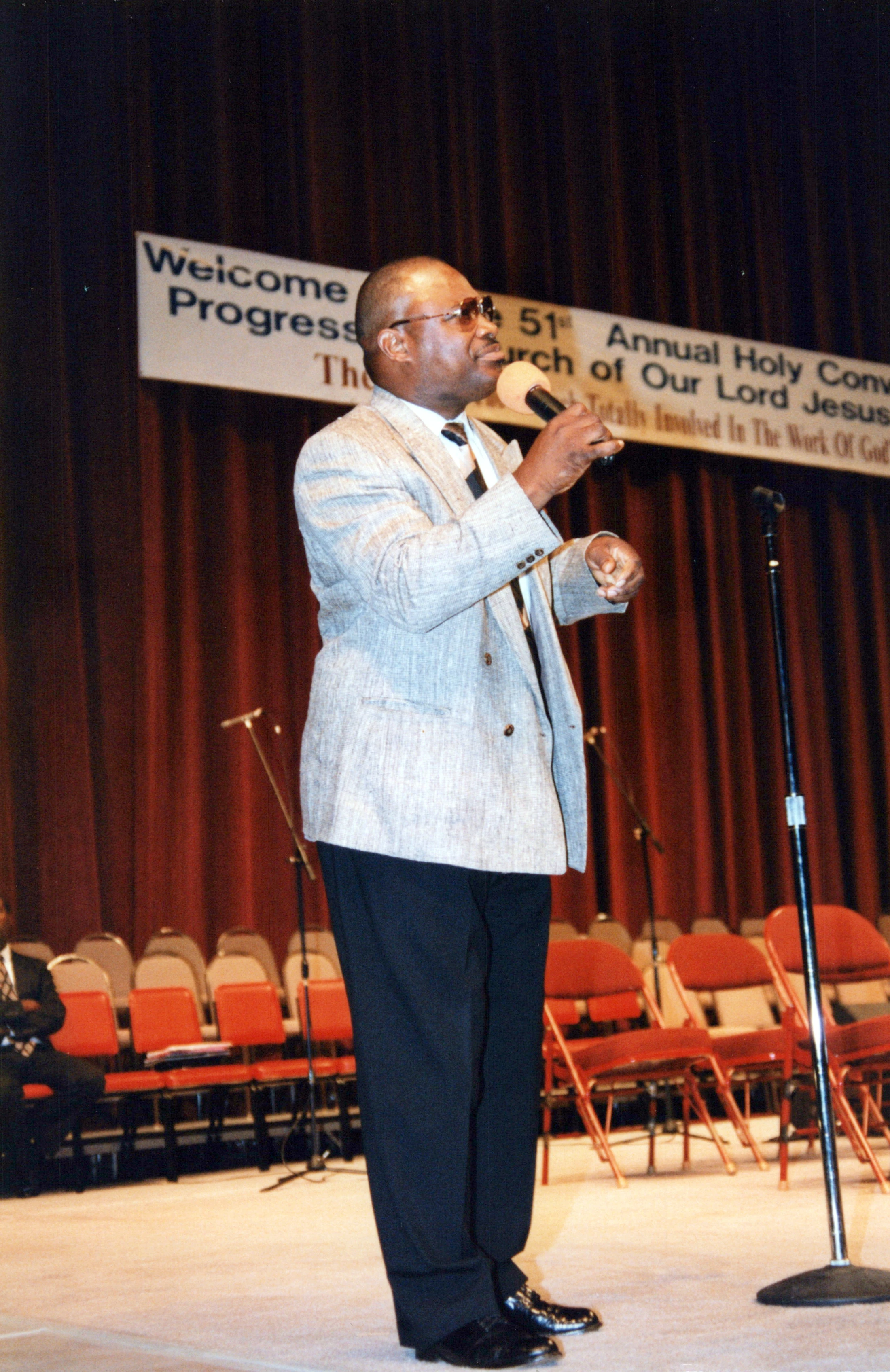 A Black man in a light grey blazer and dark pants stands on a stage speaking into a microphone during a religious convocation.