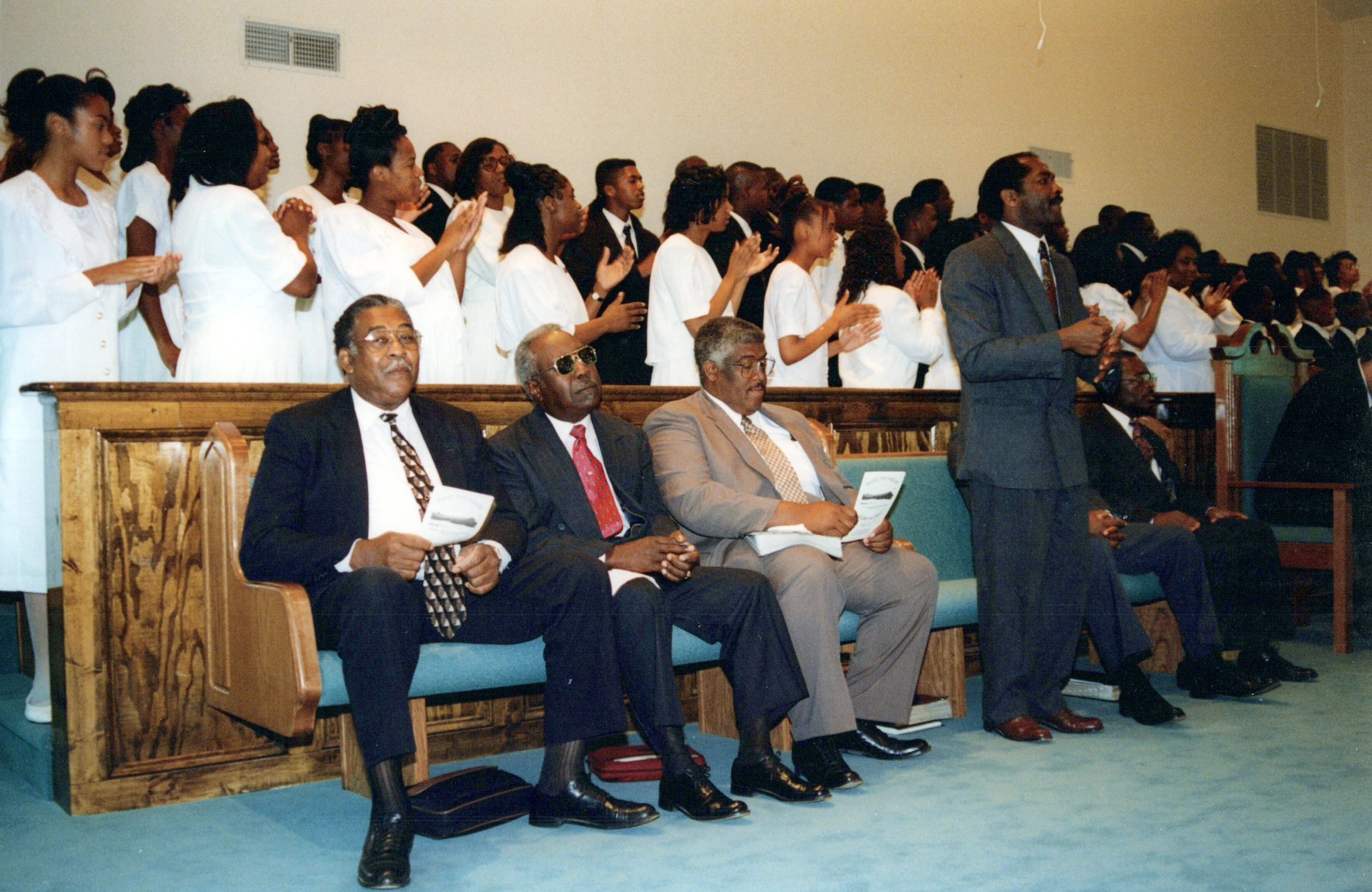 A group of older Black men in suits sit in a front church pew while a choir dressed in white stands behind them, clapping during a service.