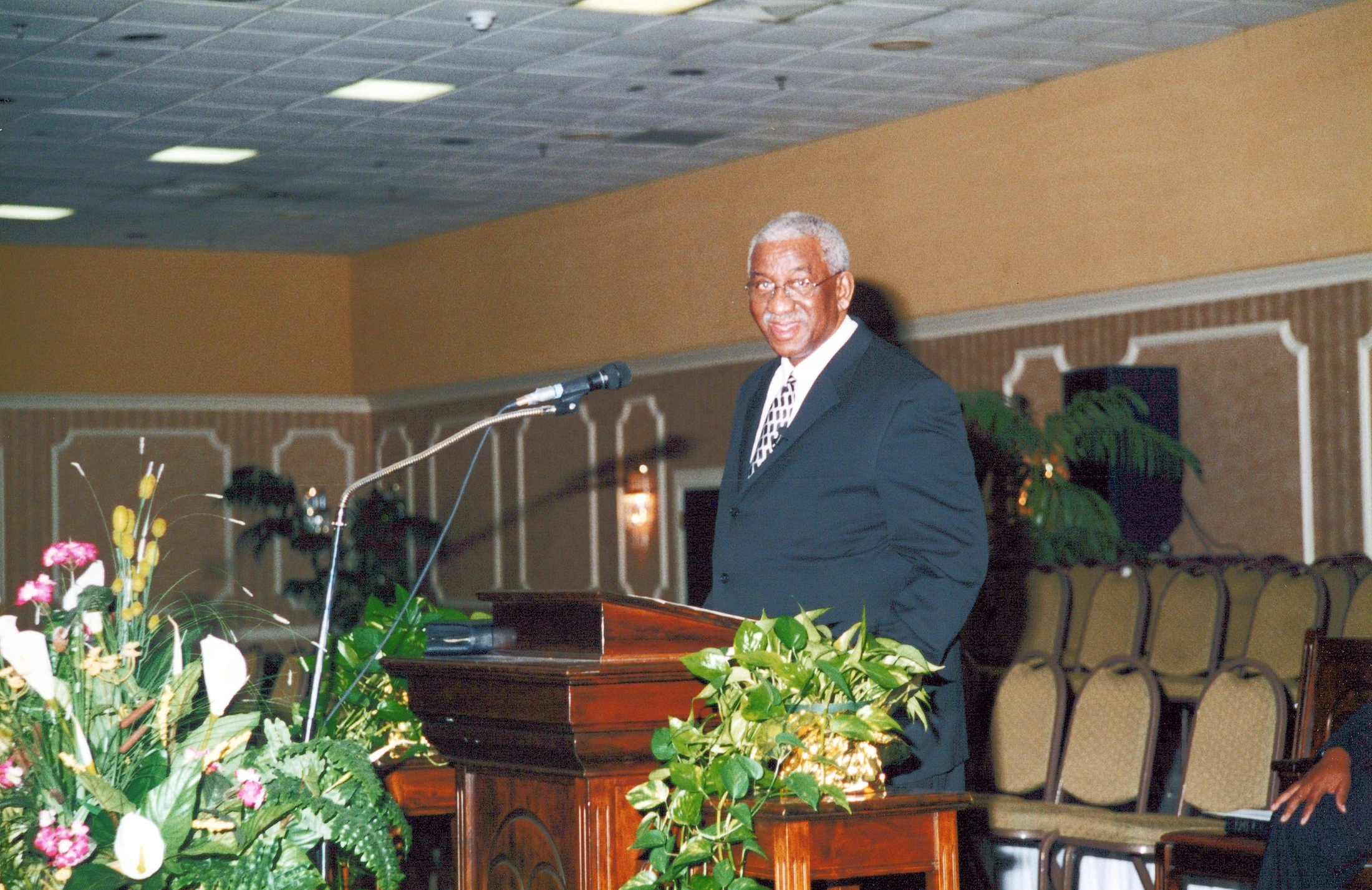 An older Black man with gray hair and glasses, wearing a dark suit, smiles while standing behind a wooden podium with a microphone in a conference room.
