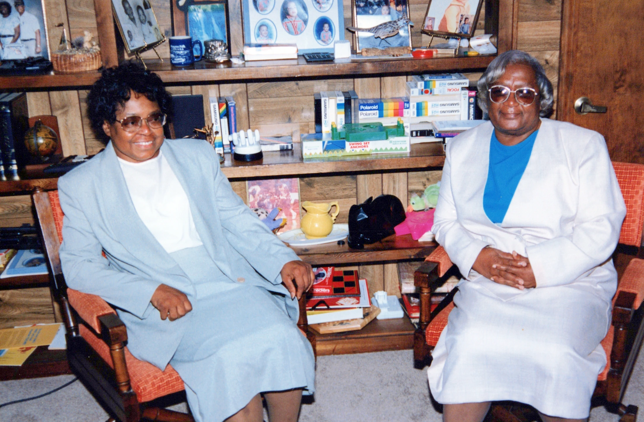Two smiling older Black women sit in chairs in front of a wood-paneled wall and bookshelf filled with family photos and mementos.