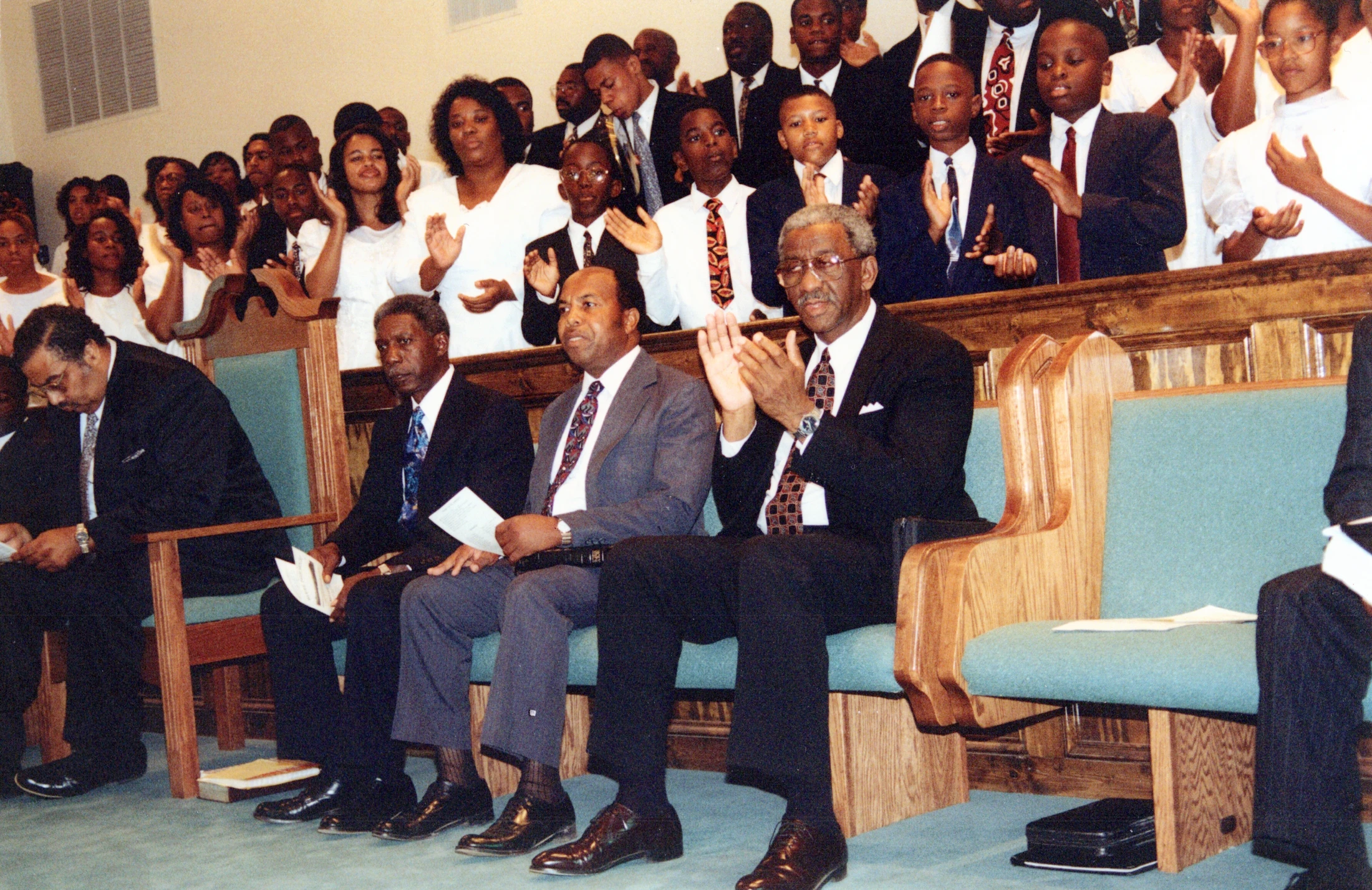 A large Black congregation, with men in suits in the front pews and women and children behind, clapping enthusiastically during a church service.