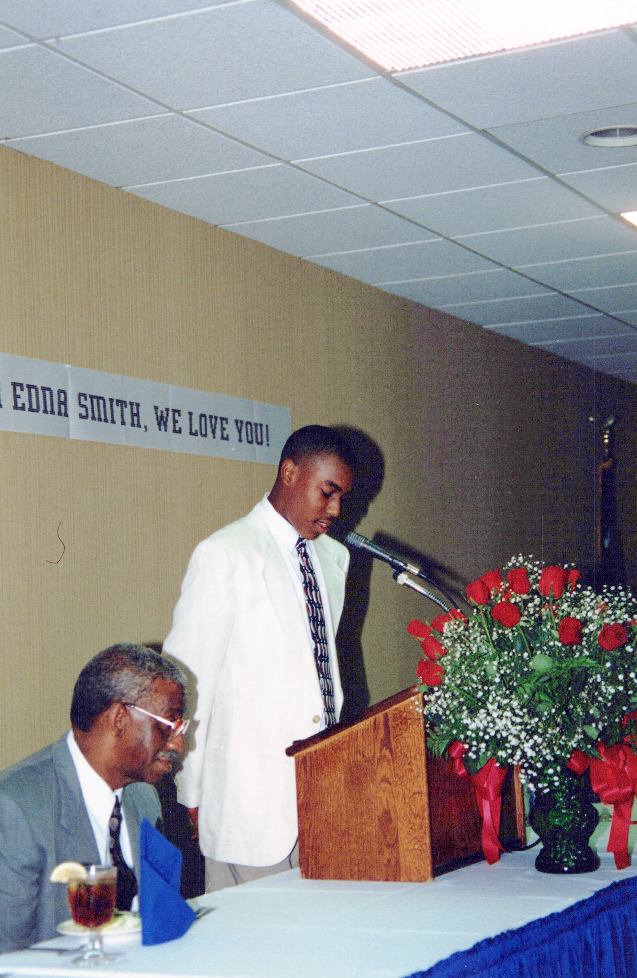 A young Black man in a white suit jacket speaks at a wooden lectern. An older man sits nearby at a table with a large bouquet of red roses.