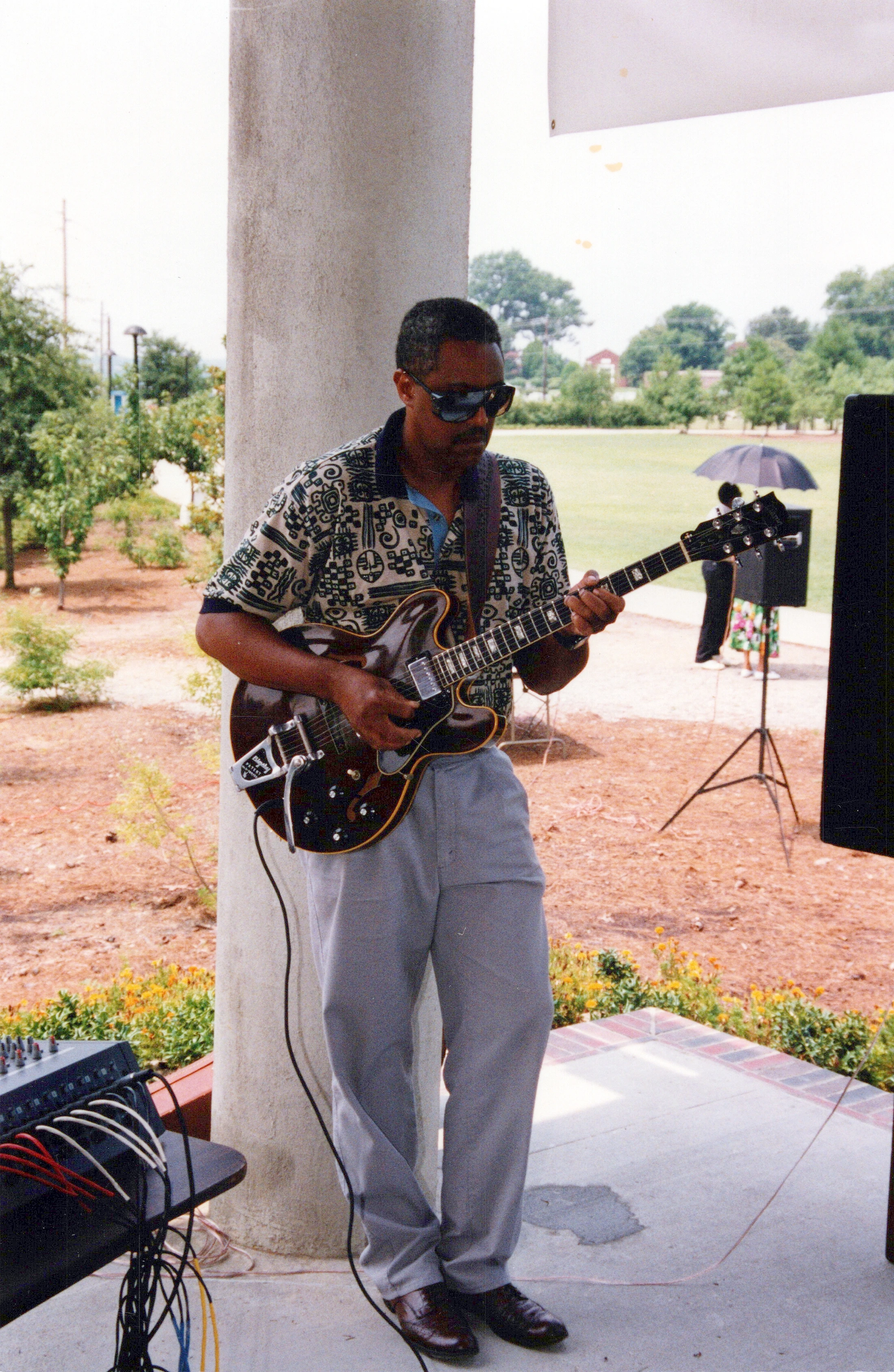 A Black man in sunglasses and a patterned shirt plays a hollow-body electric guitar while leaning against a pillar at an outdoor event.