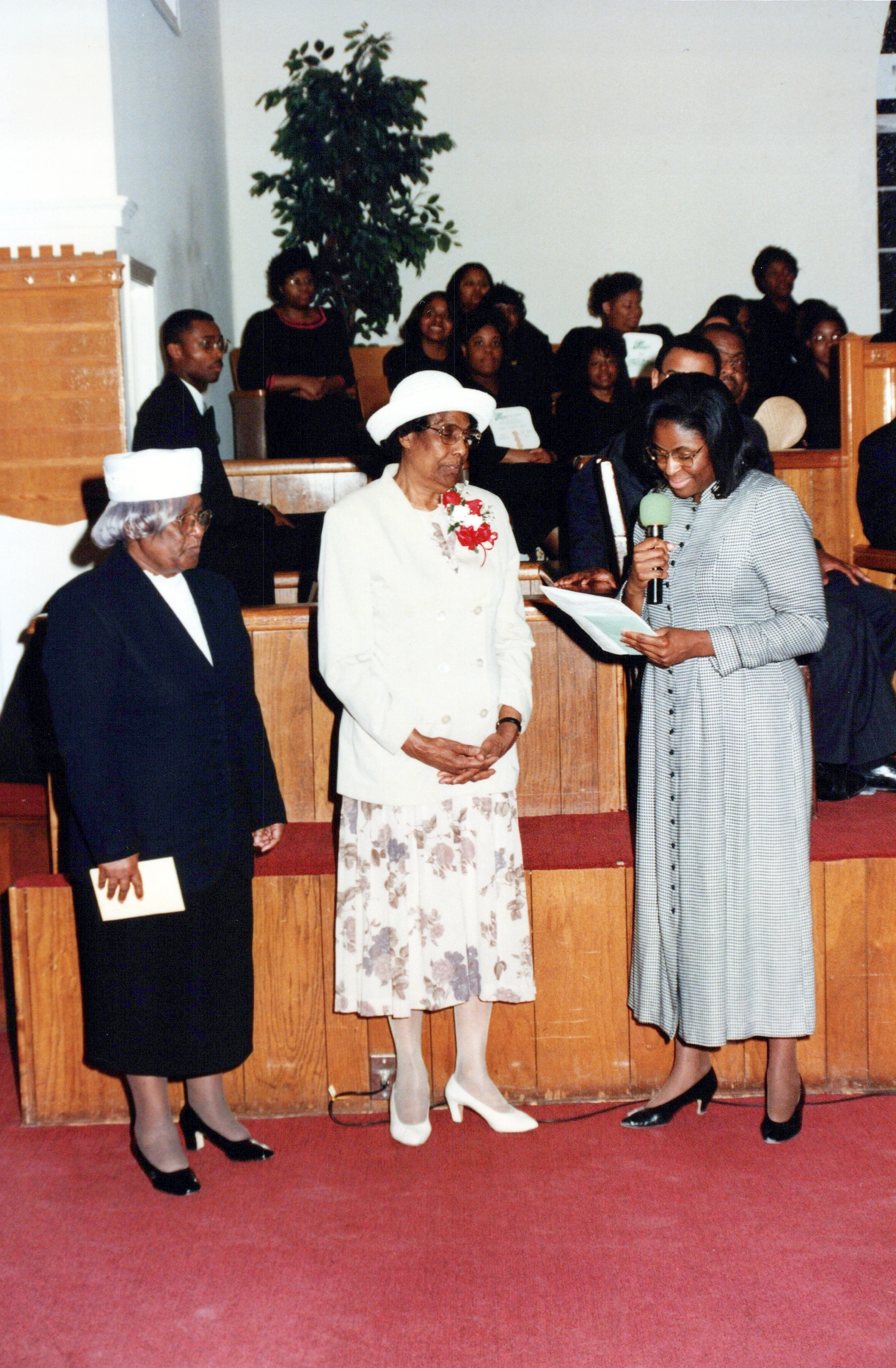 Three Black women stand at the front of a church. One woman speaks into a microphone, while the other two, wearing hats, listen attentively.