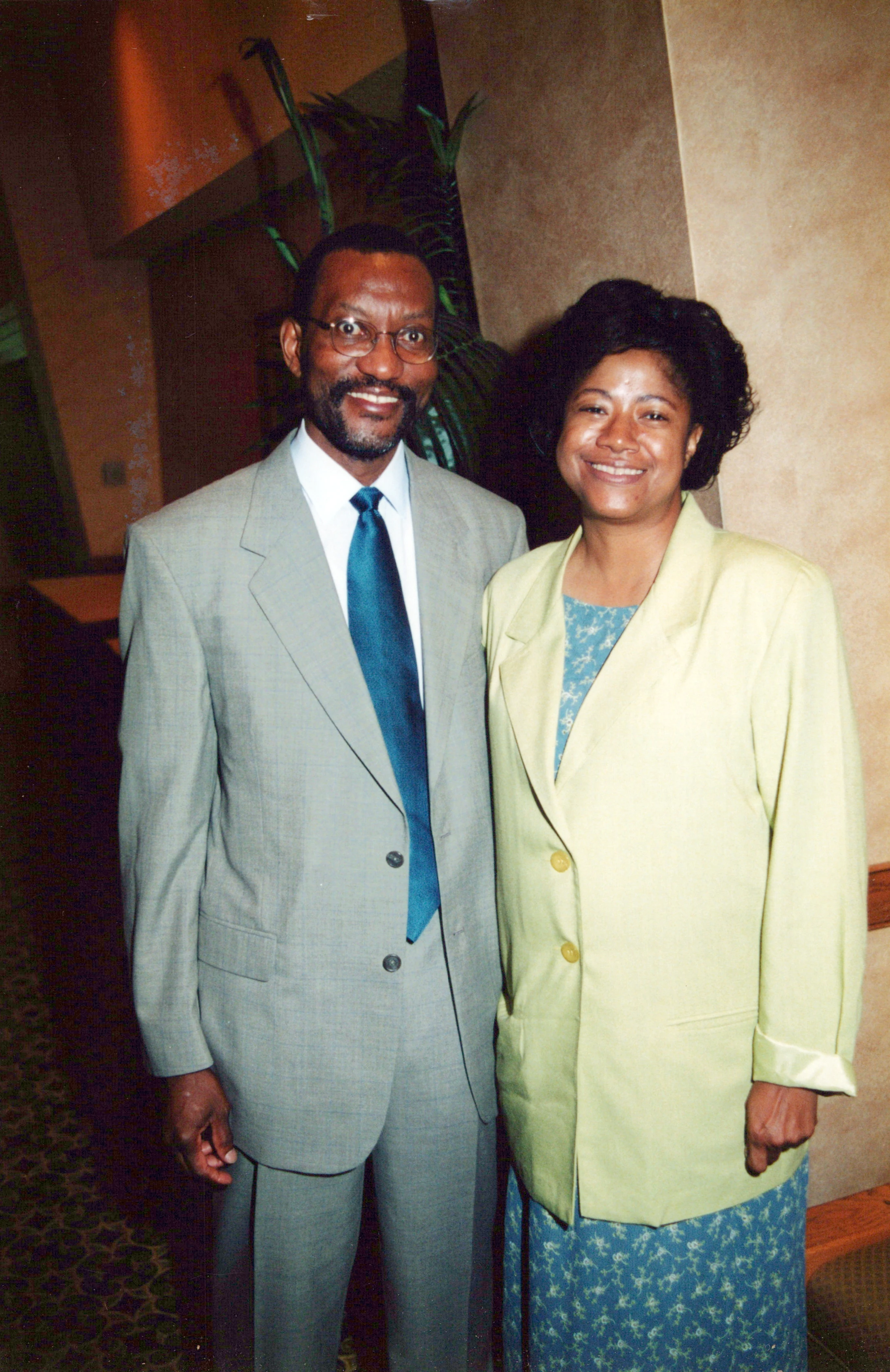 A smiling Black man in a grey suit and a woman in a light green jacket stand together for a photo indoors against a neutral wall.