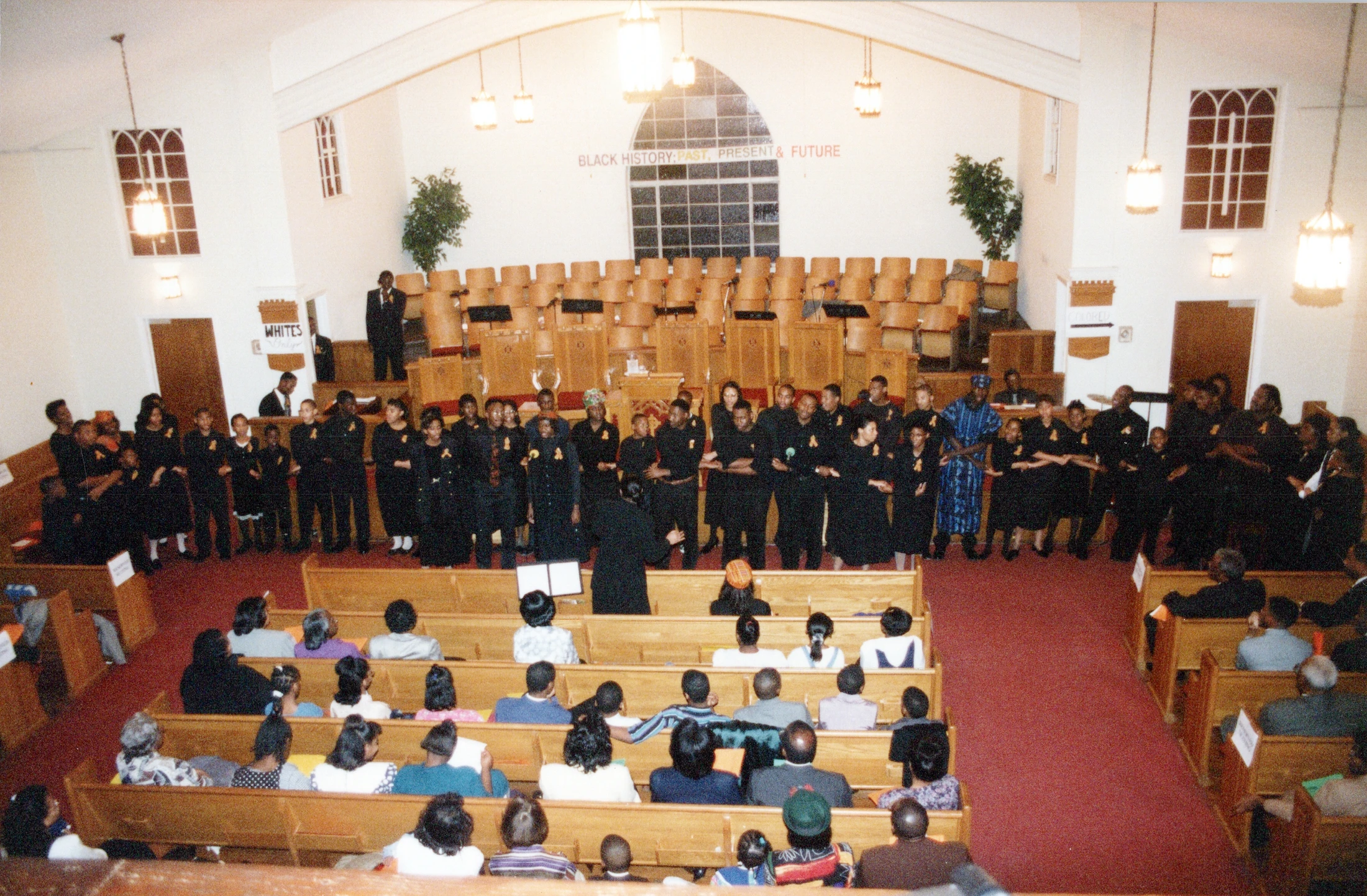 A high-angle photo of a church service. A choir stands at the front under a banner that reads "Black History: Past, Present & Future."