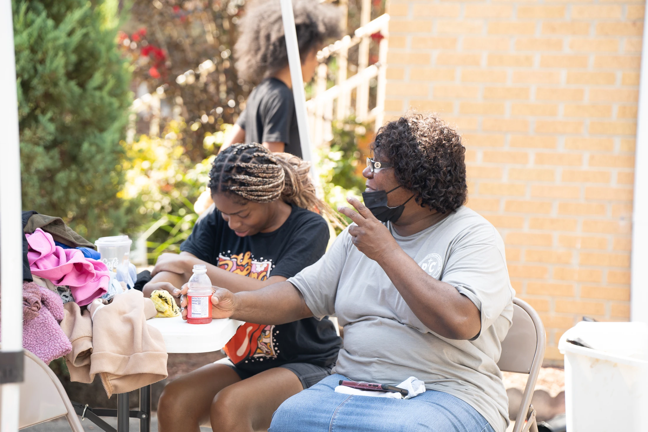 An older Black woman gestures while talking to a younger woman with braids as they sit together at a small white table outdoors on a sunny day.