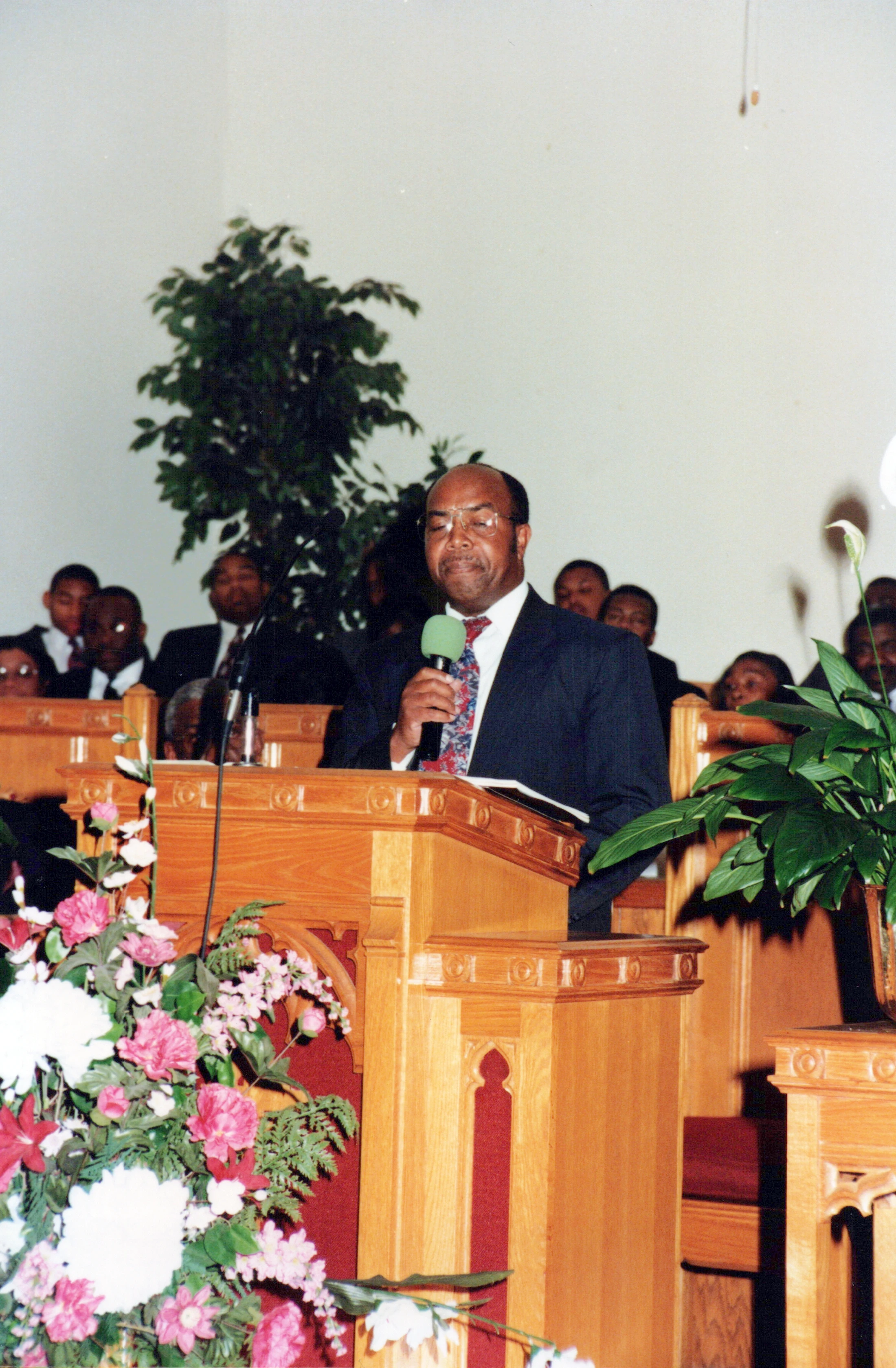 A man in a suit and glasses speaks into a microphone while standing at a wooden pulpit in a church. A congregation is seated in the background.