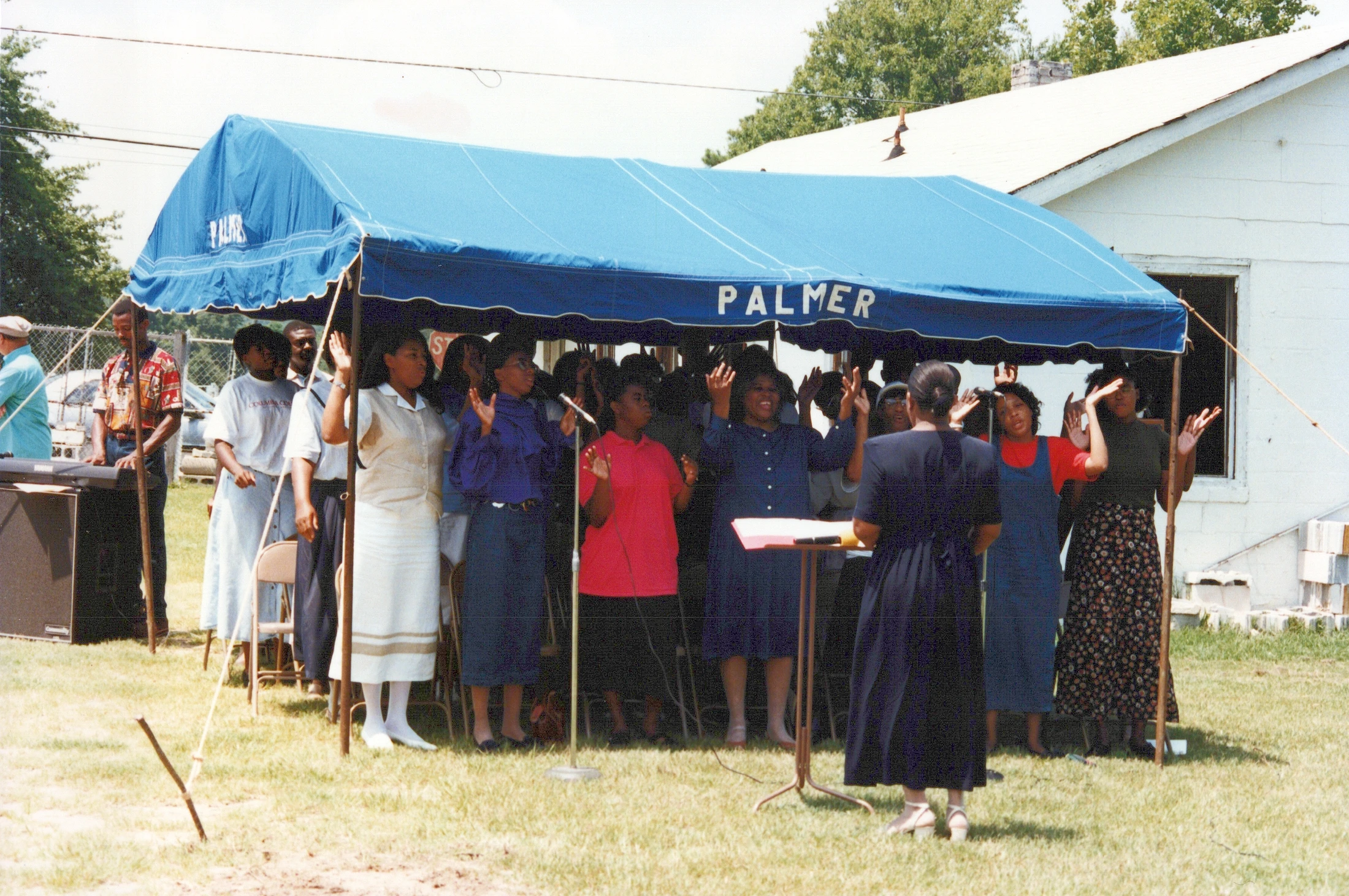 A Black choir, led by a female director, sings with raised hands under a blue tent labeled 'Palmer' during an outdoor service on a sunny day.