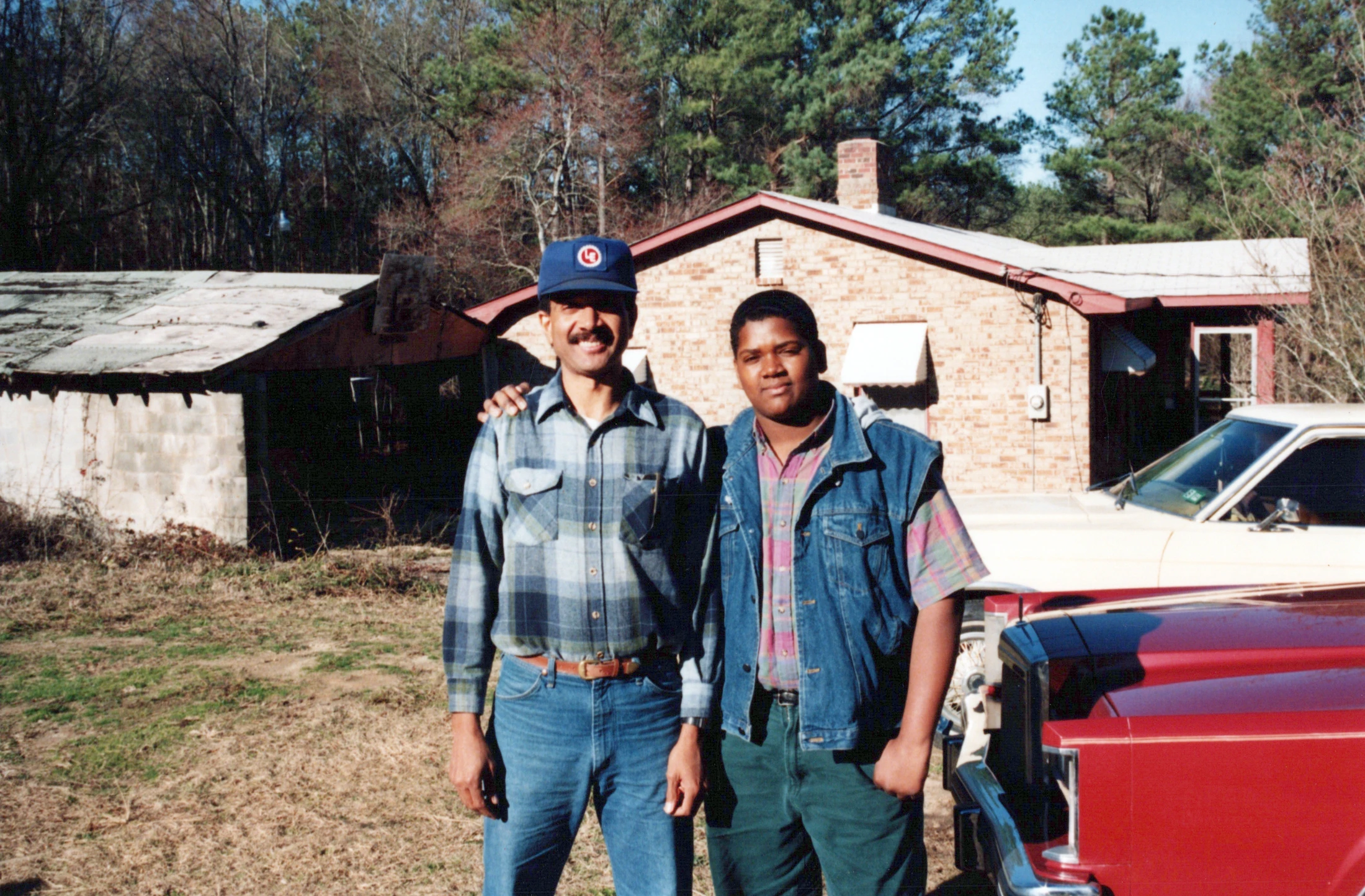 A vintage photograph of two Black men, one older and one younger, standing side-by-side outdoors with an arm around each other in front of a brick house.