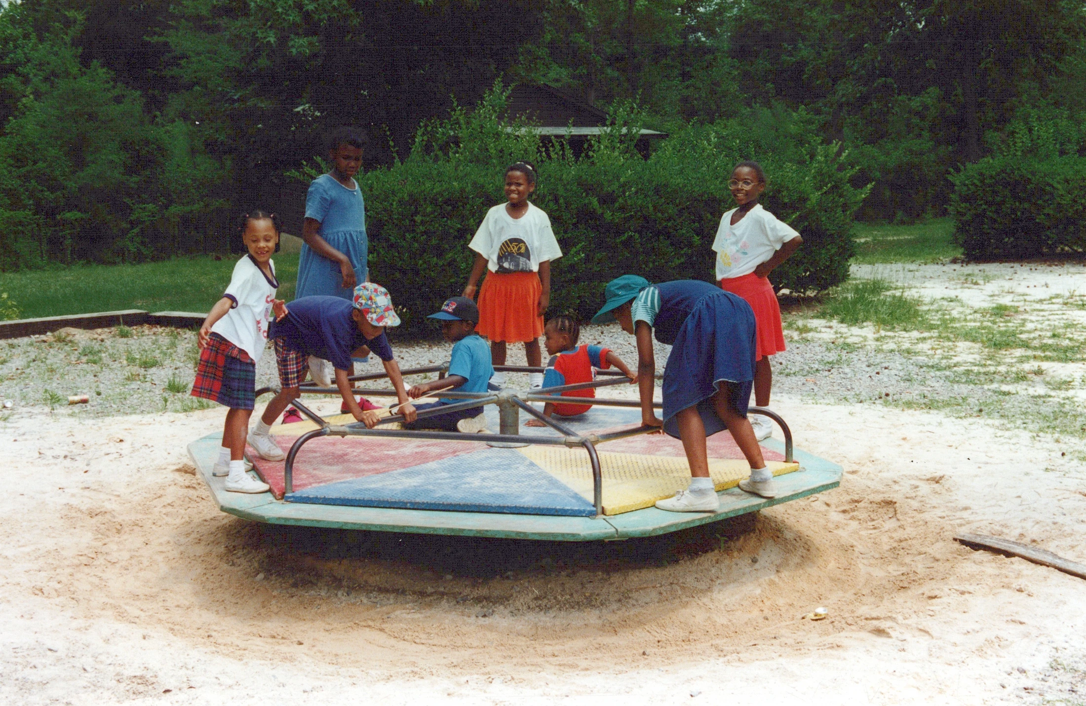 A vintage photo of seven young Black children playing on a colorful merry-go-round set in a sandy area with green trees in the background.