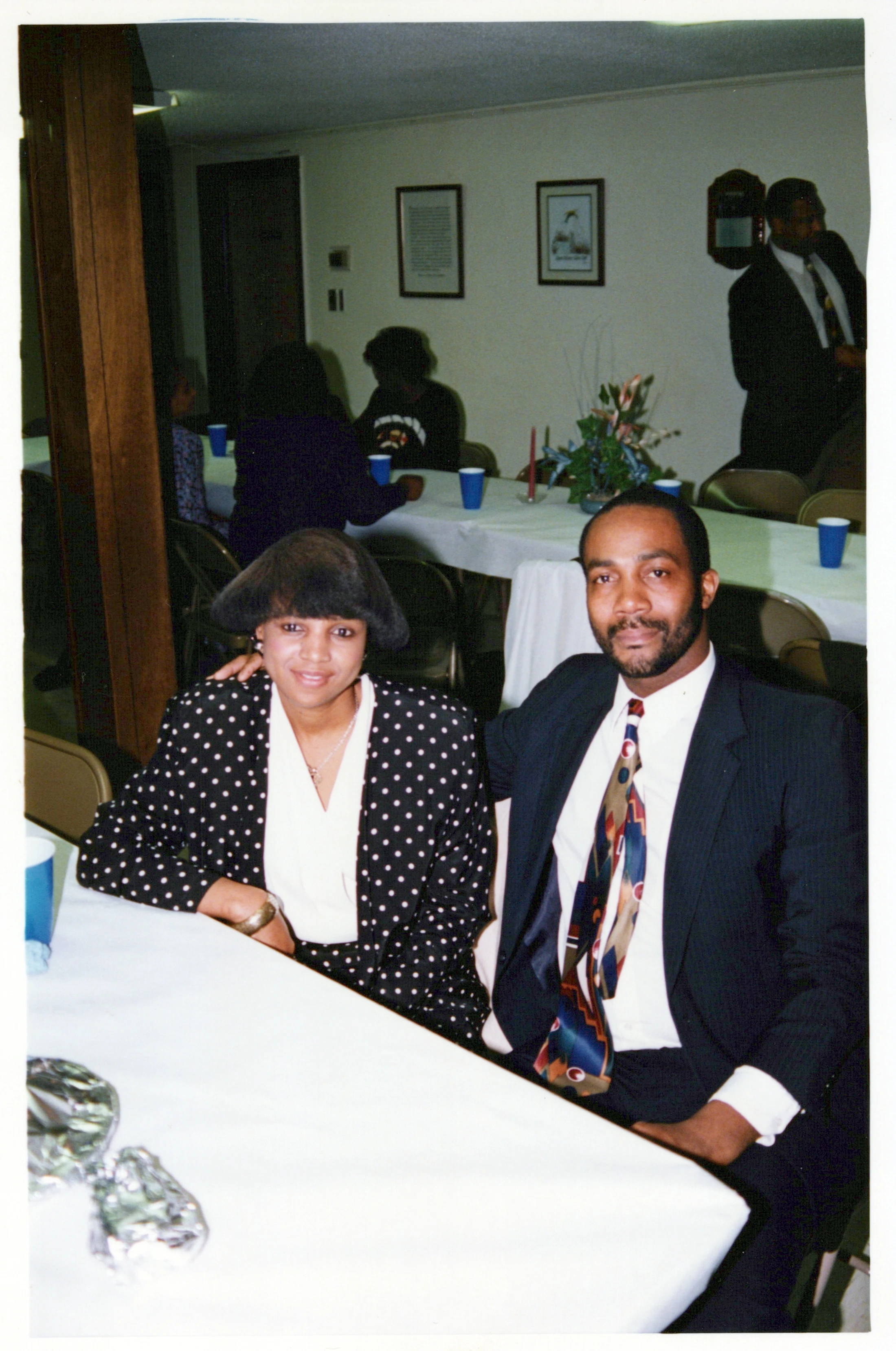 A smiling couple poses for a vintage photo. The woman wears a black and white polka dot jacket, and the man wears a dark suit with a colorful tie.