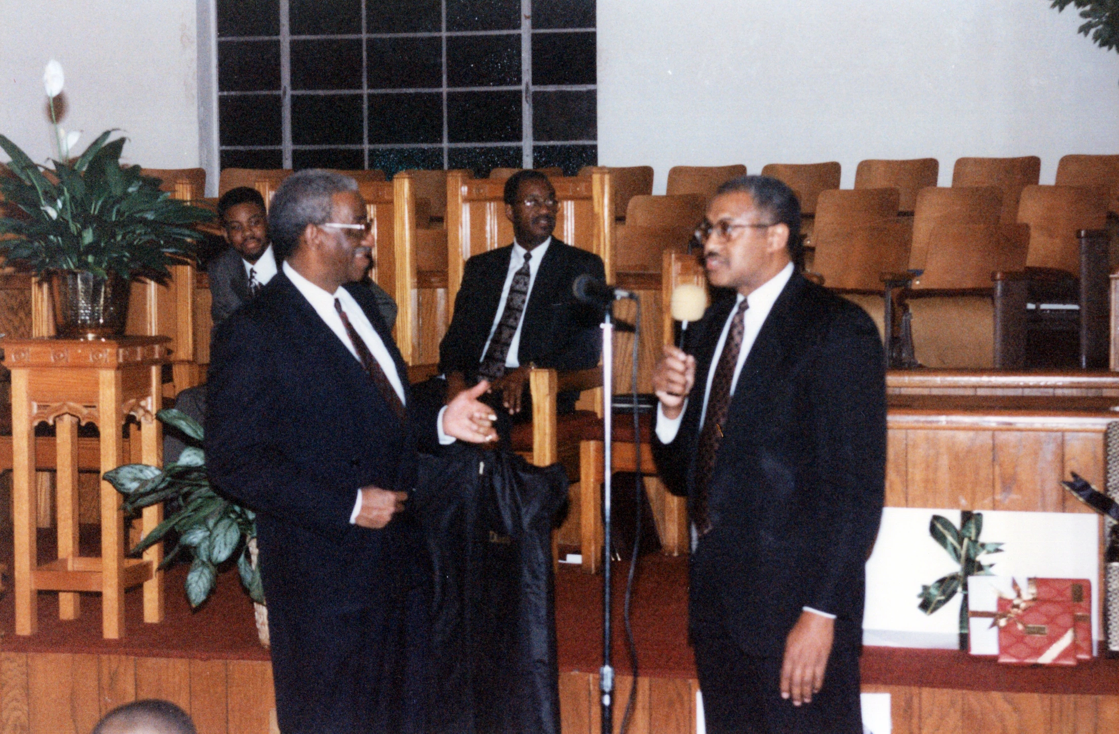 Two Black men in dark suits at the front of a church. One speaks into a microphone while the other stands beside him, gesturing. Others sit behind them.