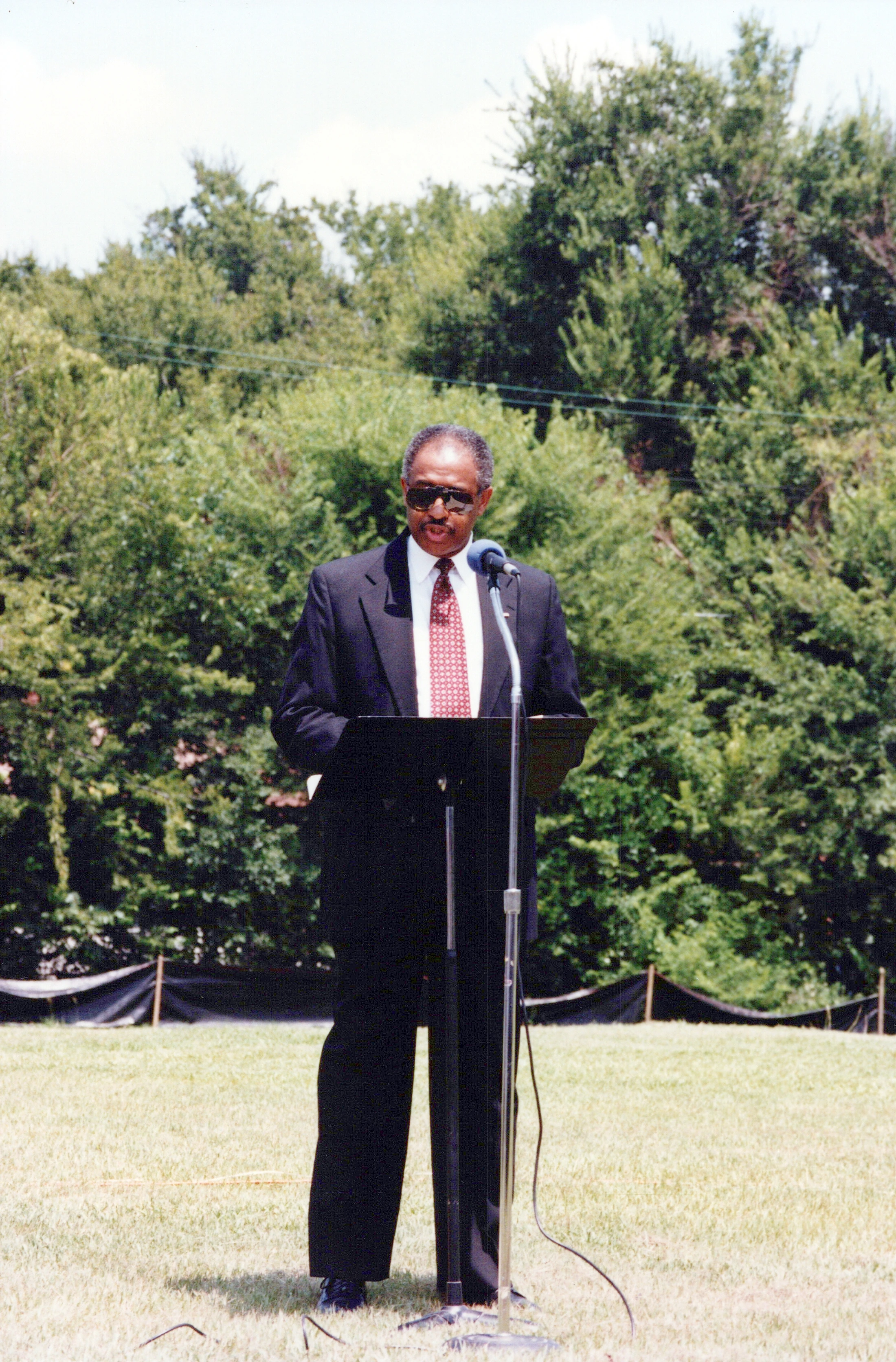 A man in a dark suit, red tie, and sunglasses stands at a lectern speaking into a microphone outdoors on a grassy field with trees in the background.