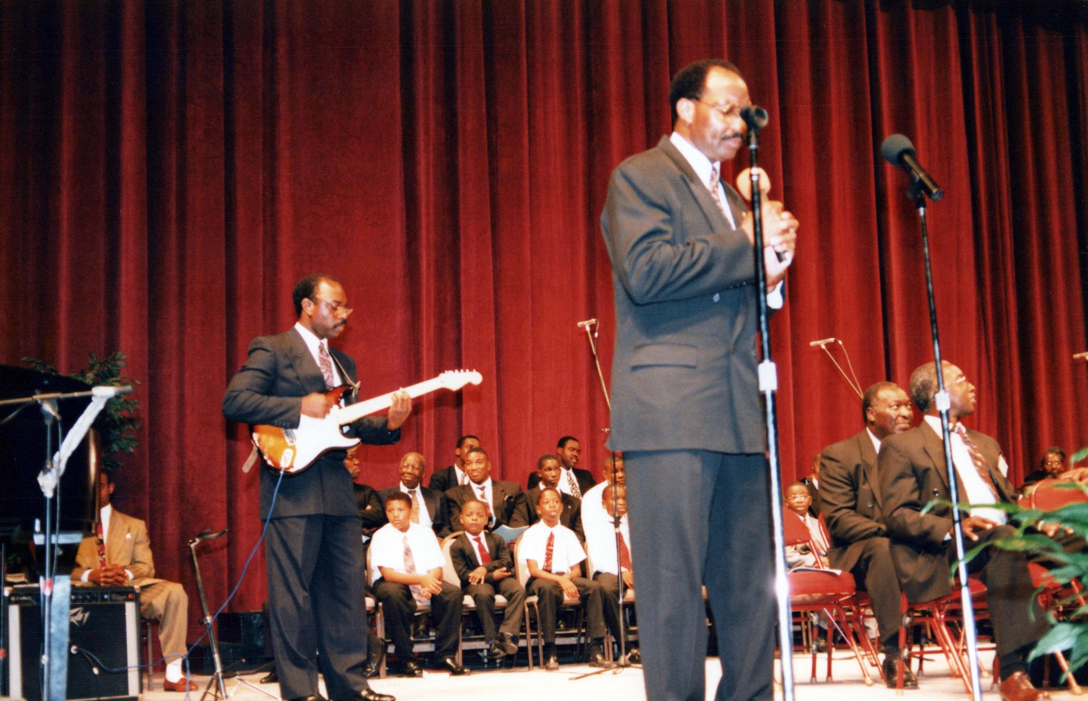 A Black man sings into a microphone on a stage while another man plays an electric guitar. In the background, a choir of men and boys watches from their seats.