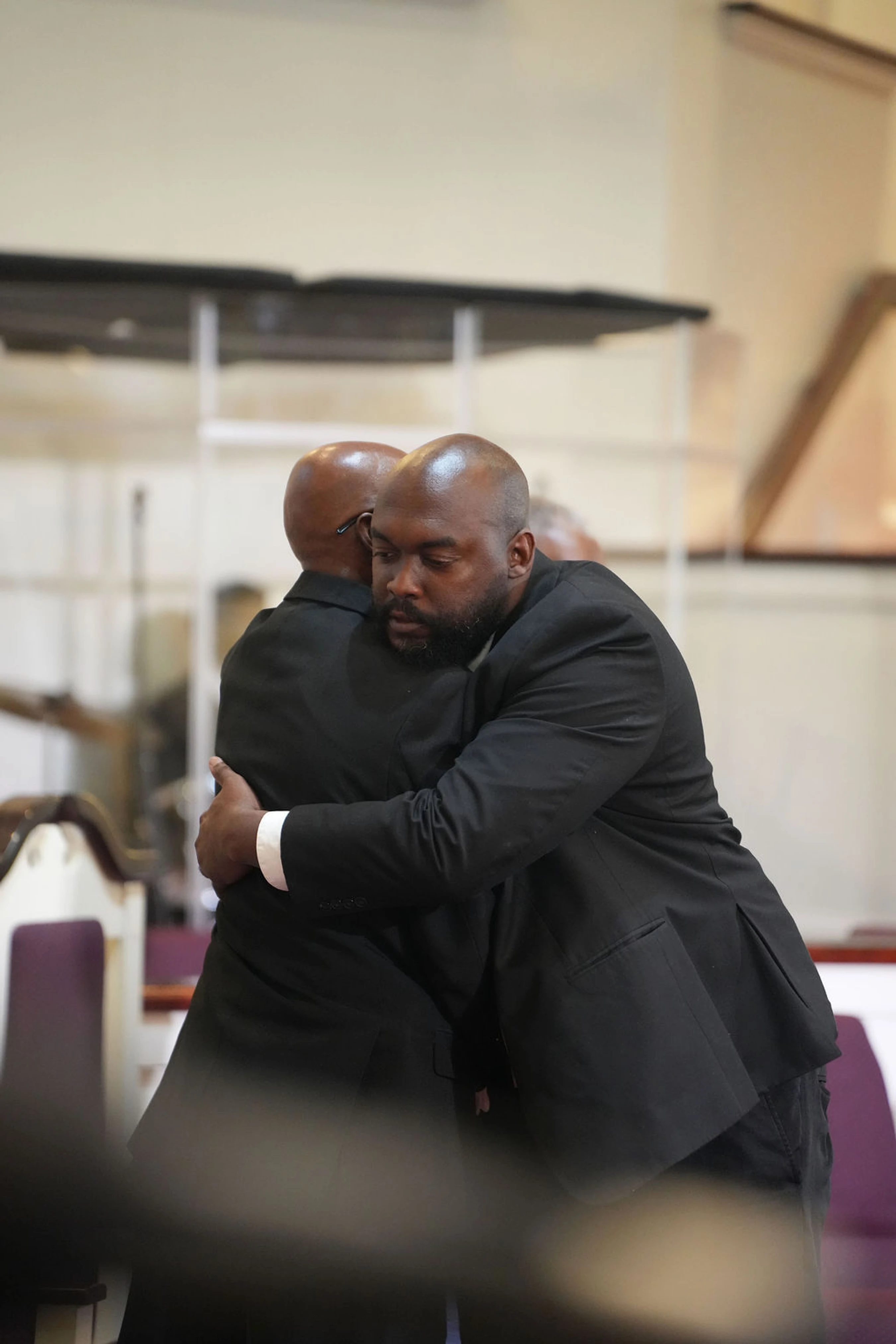 Two bald Black men in black suits share a solemn embrace inside what appears to be a church, offering comfort to one another.