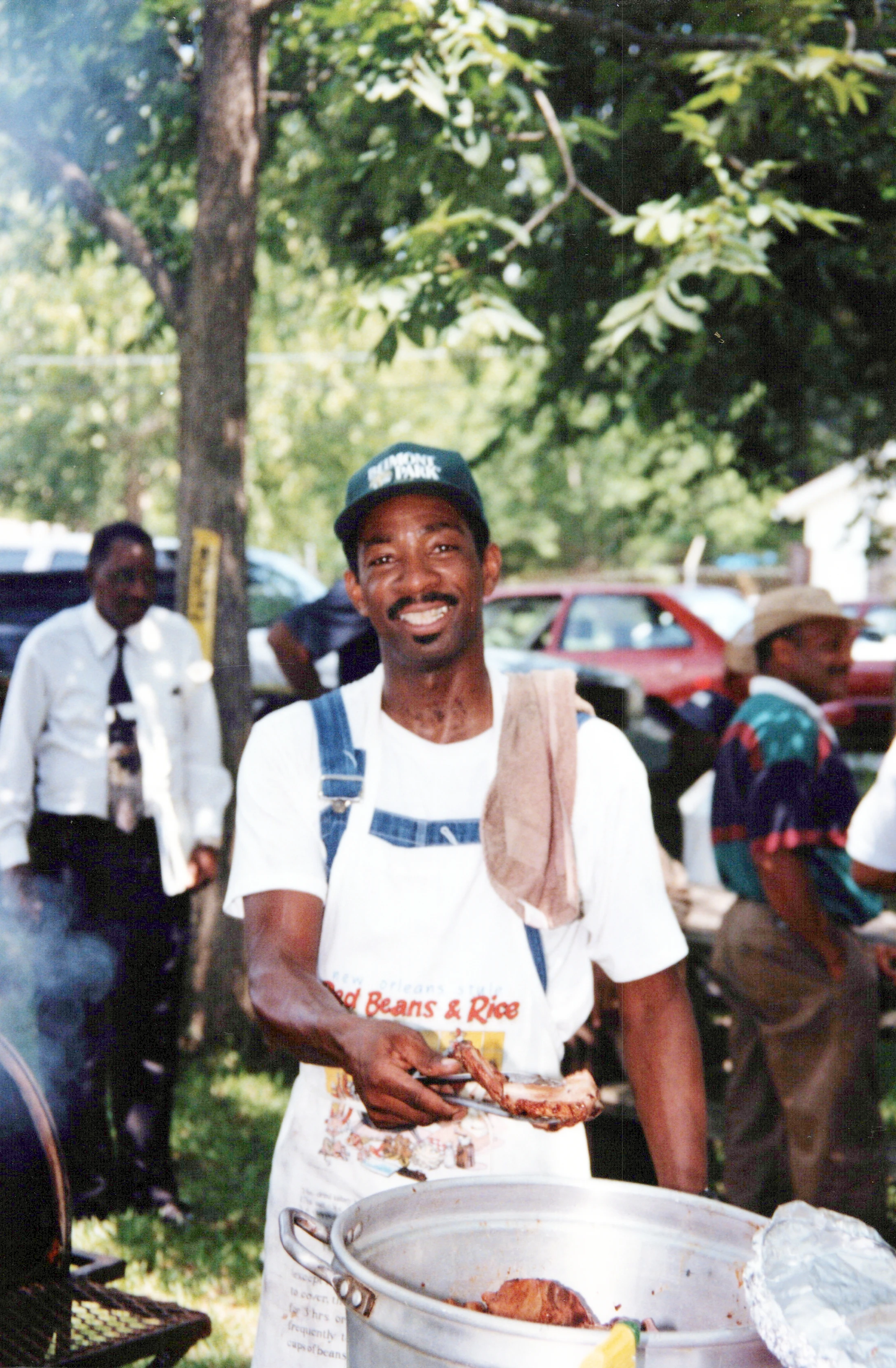 A smiling Black man in an apron and overalls holds up a piece of meat with tongs over a large pot while cooking at an outdoor barbecue.