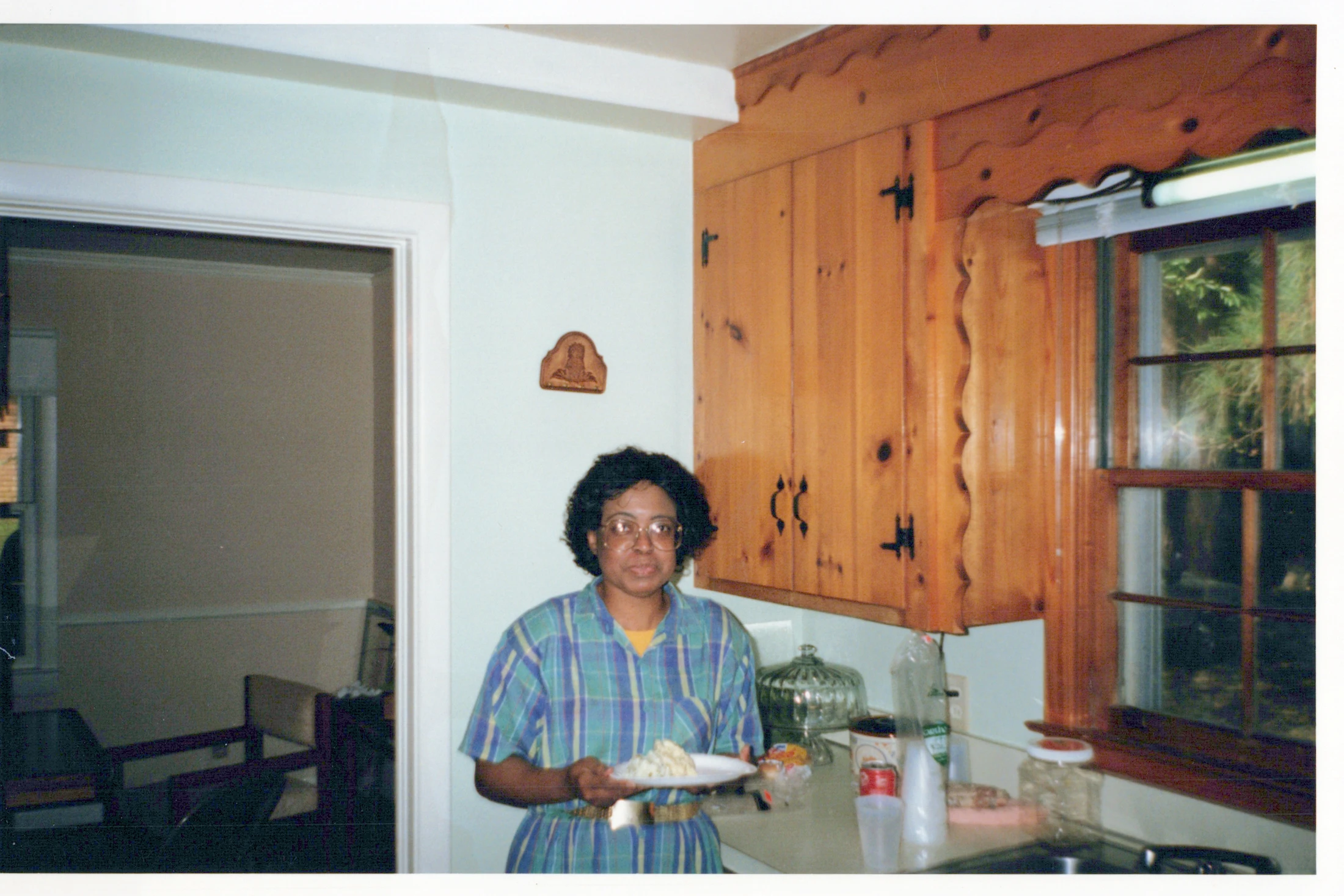 A vintage photograph of a Black woman with glasses and a plaid dress standing in a kitchen. She is holding a white plate with food and looking at the camera.