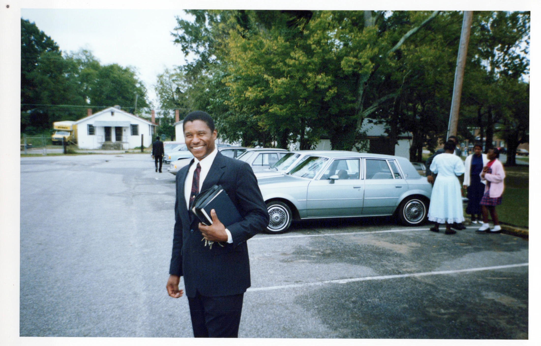 A smiling Black man in a dark suit holds a book and keys while standing in a parking lot. Other people, cars, and a small building are in the background.