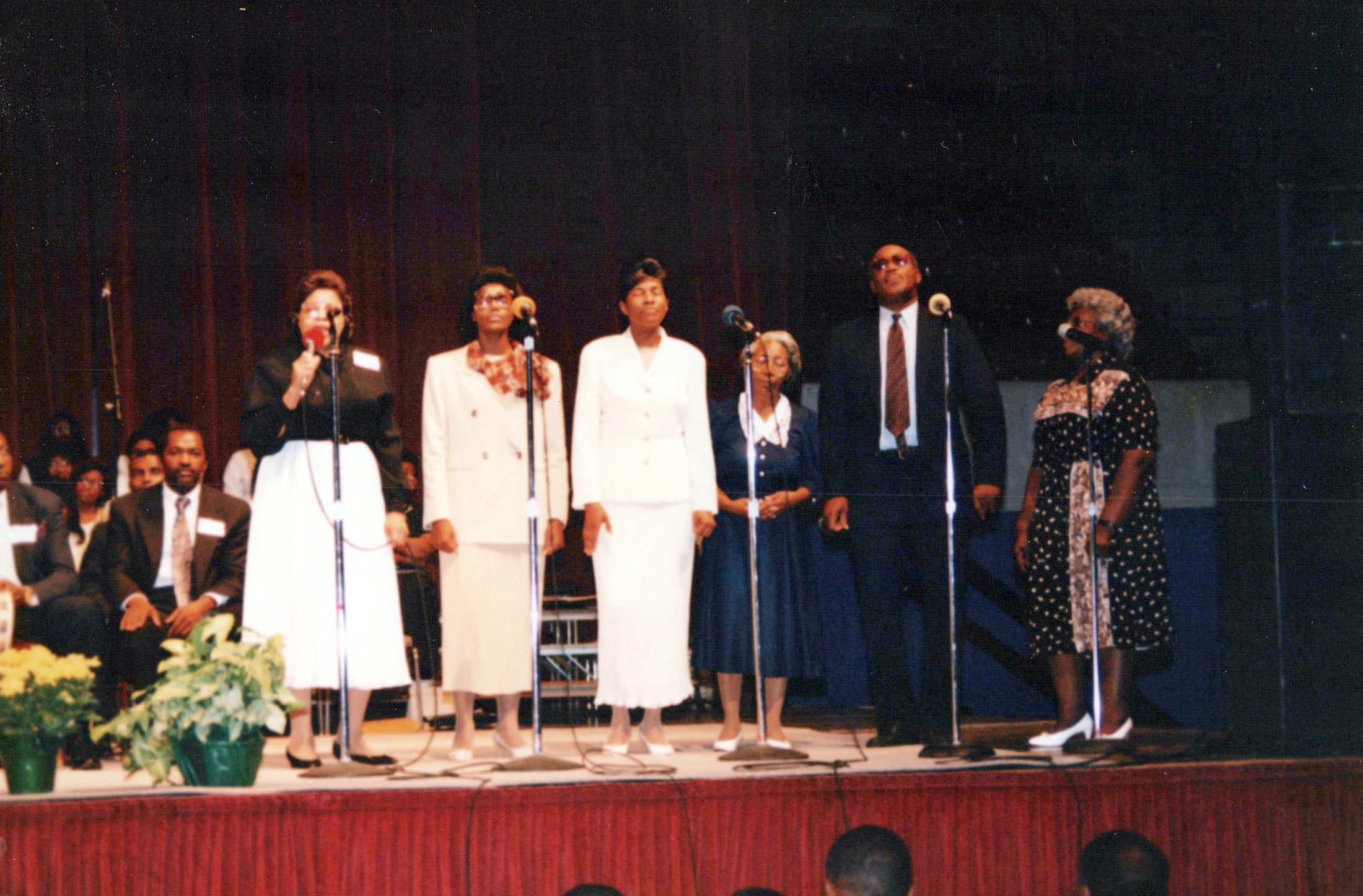 A grainy, vintage photo of six African American singers, five women and one man, standing in a line on a stage in front of microphones during a performance.