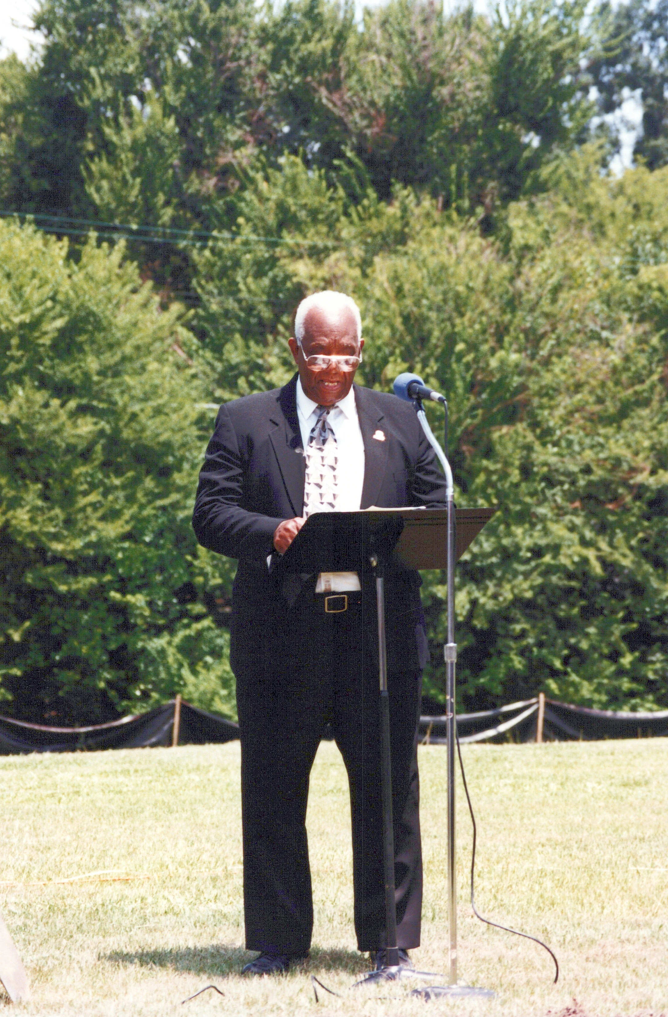 An older Black man with white hair and glasses, wearing a dark suit, speaks at a lectern with a microphone in a grassy field with trees behind him.