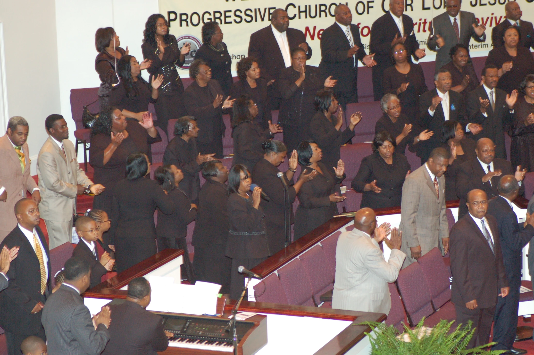 A large African American gospel choir and congregation stand, sing, and clap their hands joyfully during a worship service in their church.