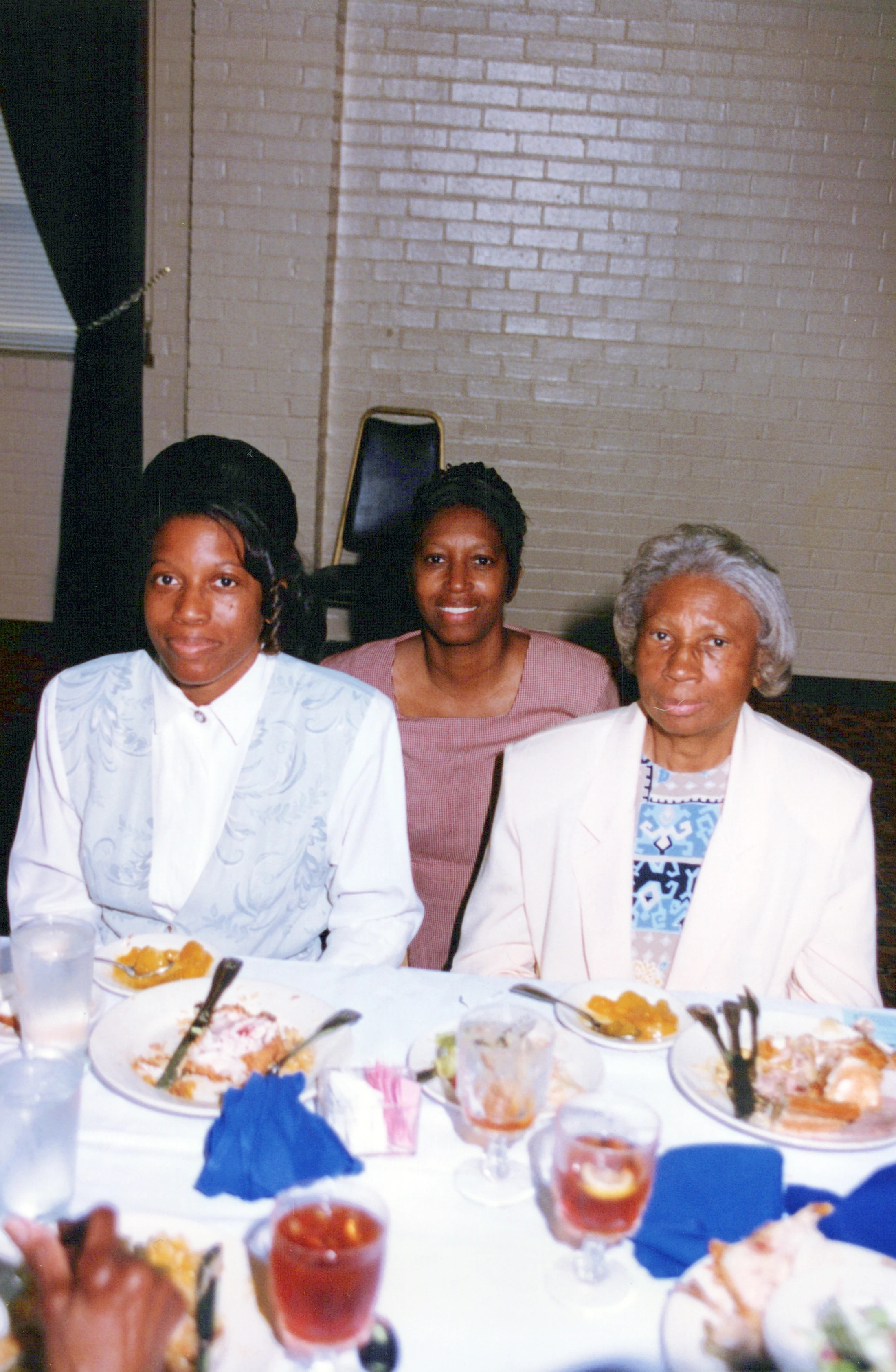 A vintage photograph of three Black women of different ages seated together at a dinner table, posing for the camera with plates of food in front of them.