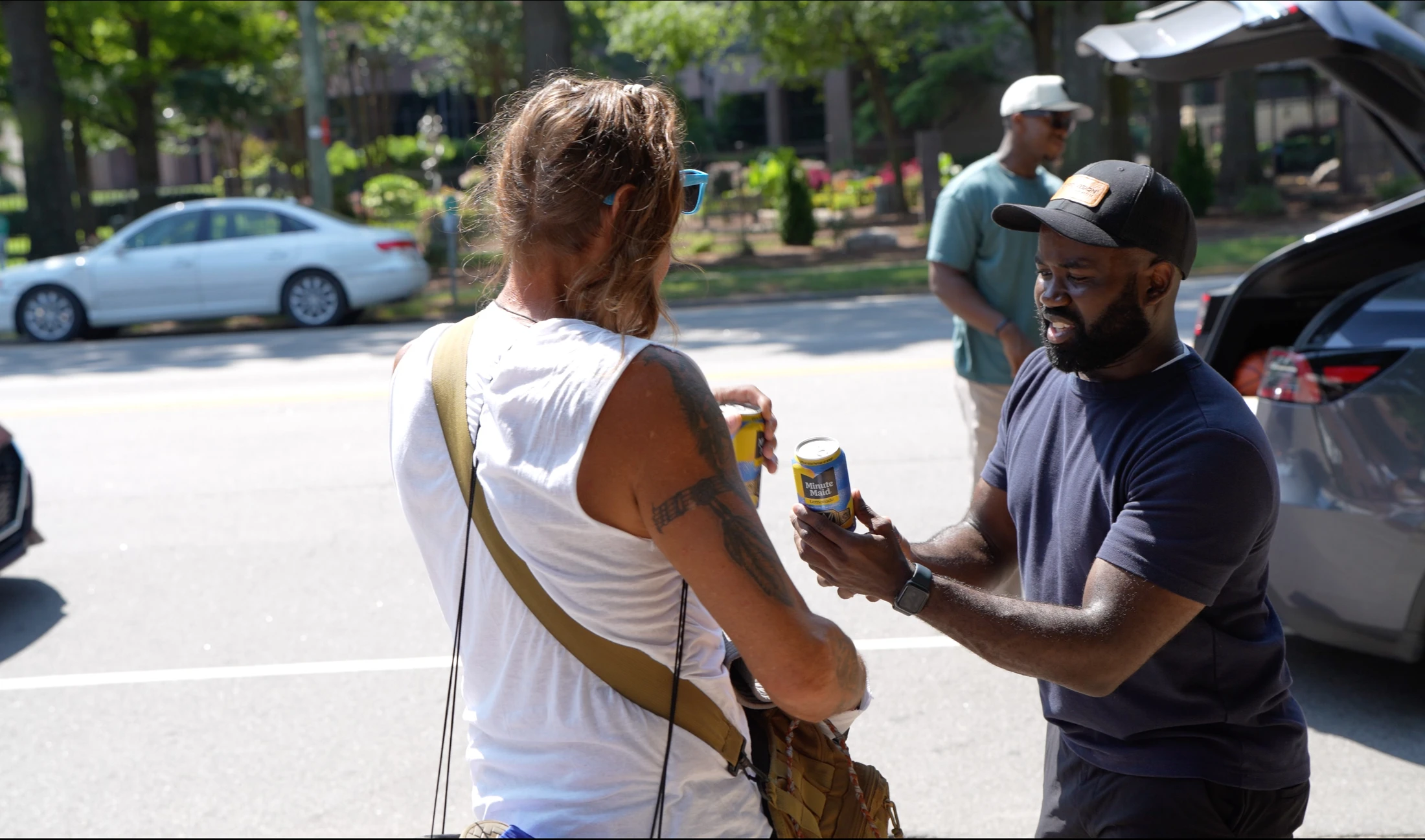 A man in a blue shirt and baseball cap smiles as he hands a can of lemonade to another person on a sunny street next to a car with its trunk open.