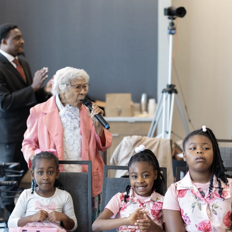 An elderly Black woman in a pink jacket speaks into a microphone while three young girls sit in the front row, listening at an event.