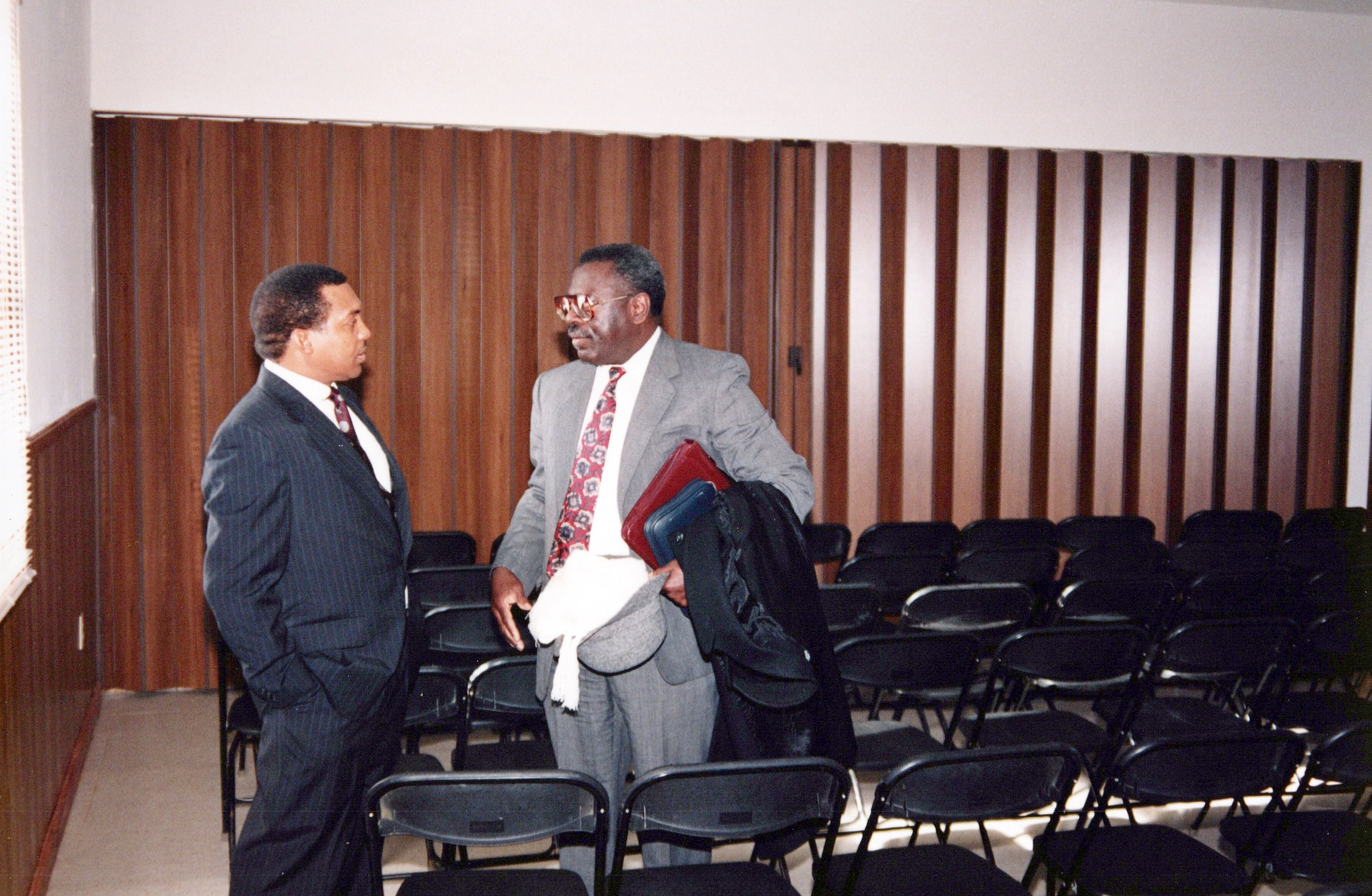 Two African American men in business suits stand talking in a room filled with empty black folding chairs. The room has vertical wood paneling on the walls.