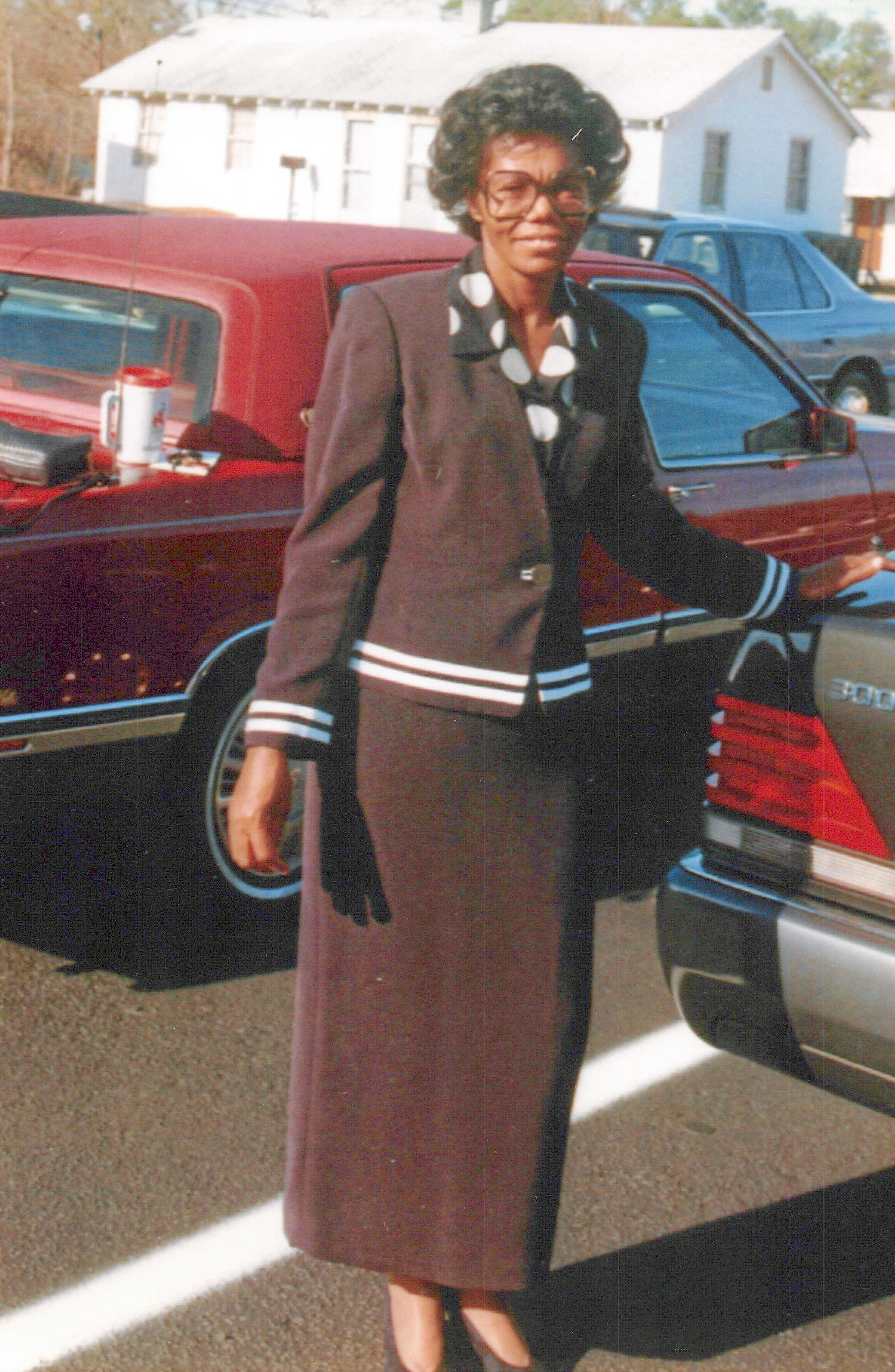 A vintage, full-length photo of an African American woman wearing glasses and a dark suit with white trim, standing between two cars in a sunny parking lot.