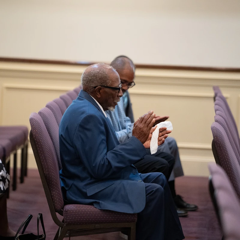 Side profile of an elderly Black man in a blue suit sitting in a purple church pew. He holds a tissue and has his hands clasped together in contemplation.