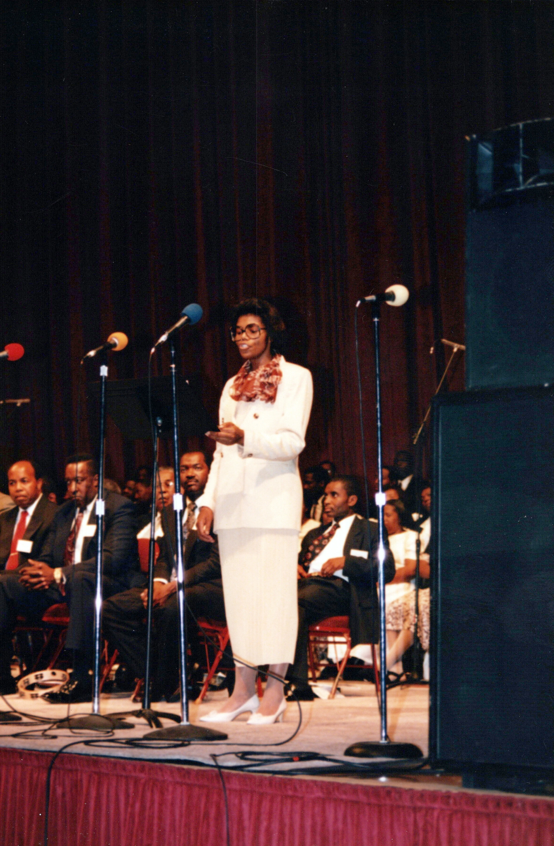 A Black woman in a white suit and glasses stands on a stage speaking into several microphones. An audience is seated behind her.