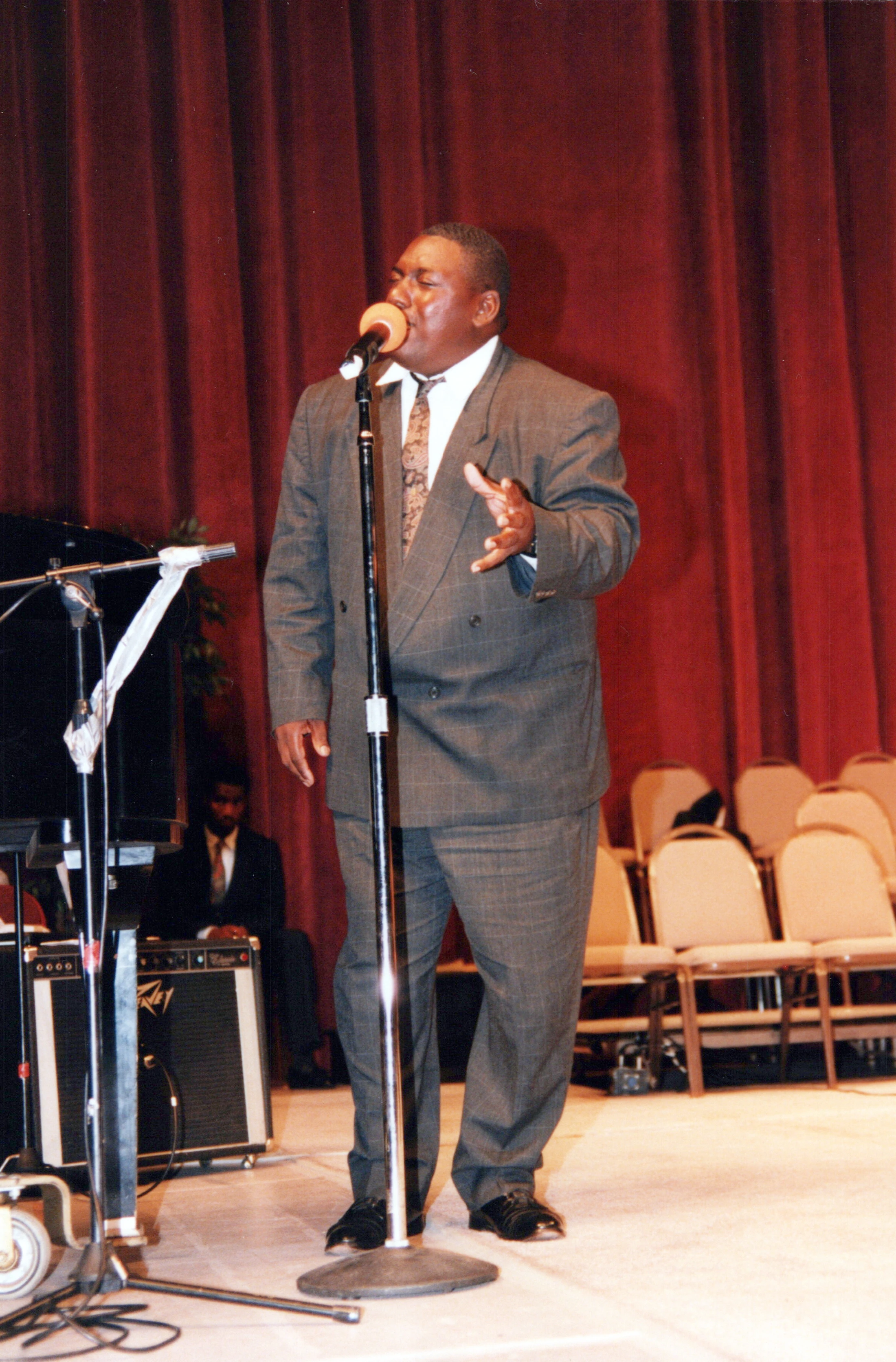 A man in a gray plaid suit sings passionately into a microphone on stage, with his eyes closed and one hand raised, in front of a red velvet curtain.