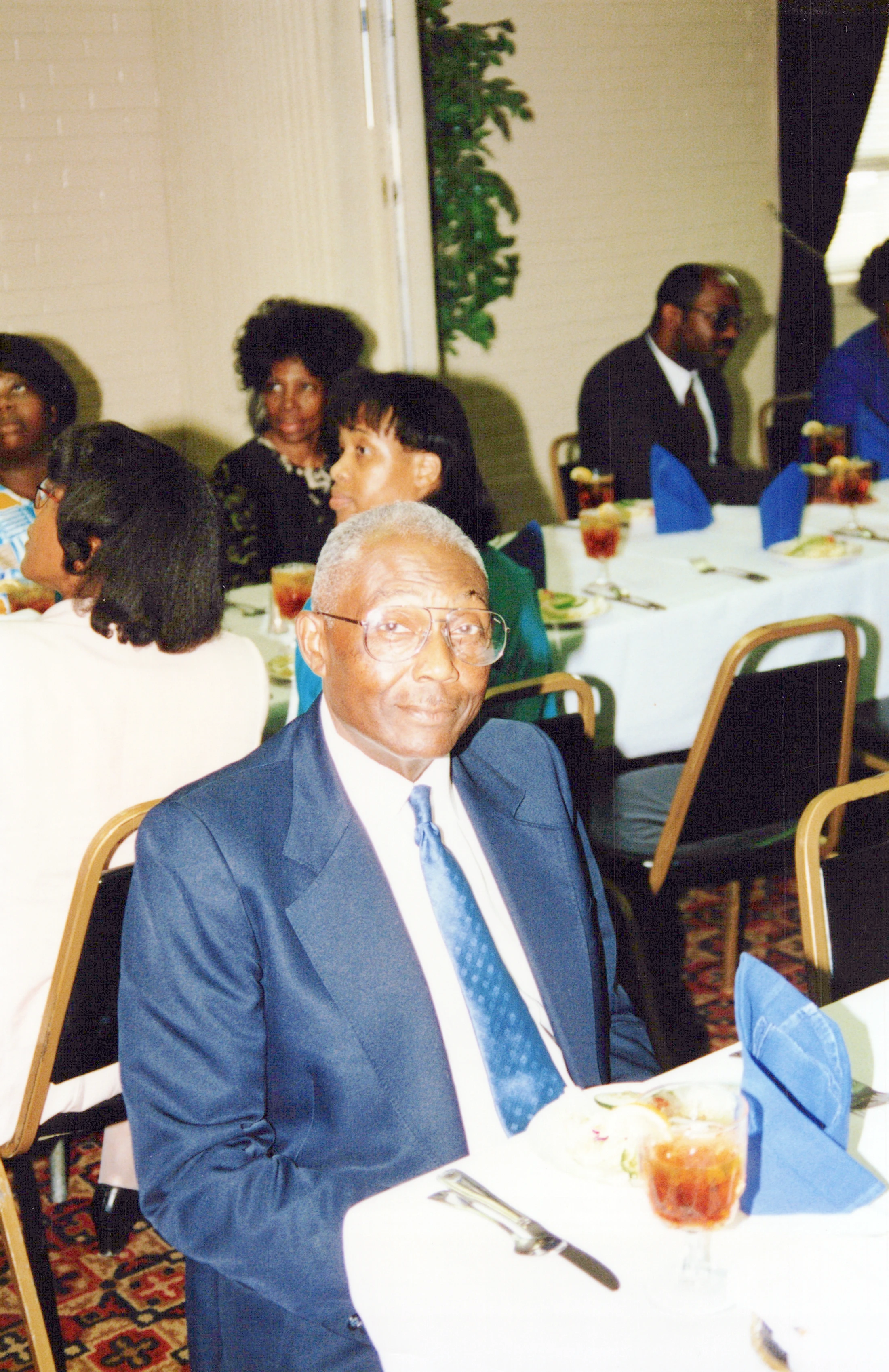 An older Black man in a blue suit and glasses smiles at the camera while seated at a dining table during a formal event with other guests in the background.