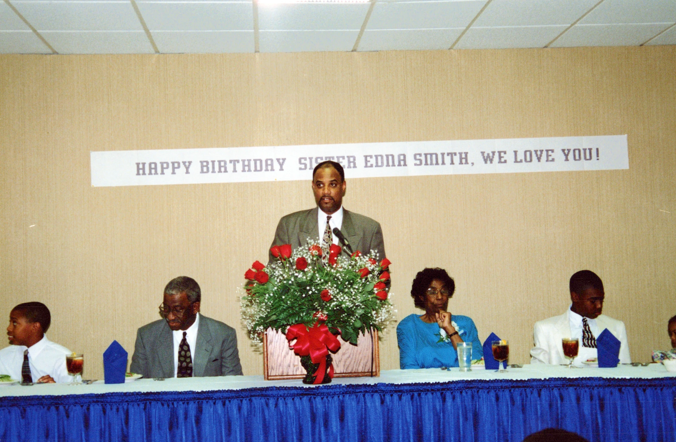 A man in a suit speaks at a podium in front of a banner that reads "Happy Birthday Sister Edna Smith, We Love You!" with guests seated at a long table.