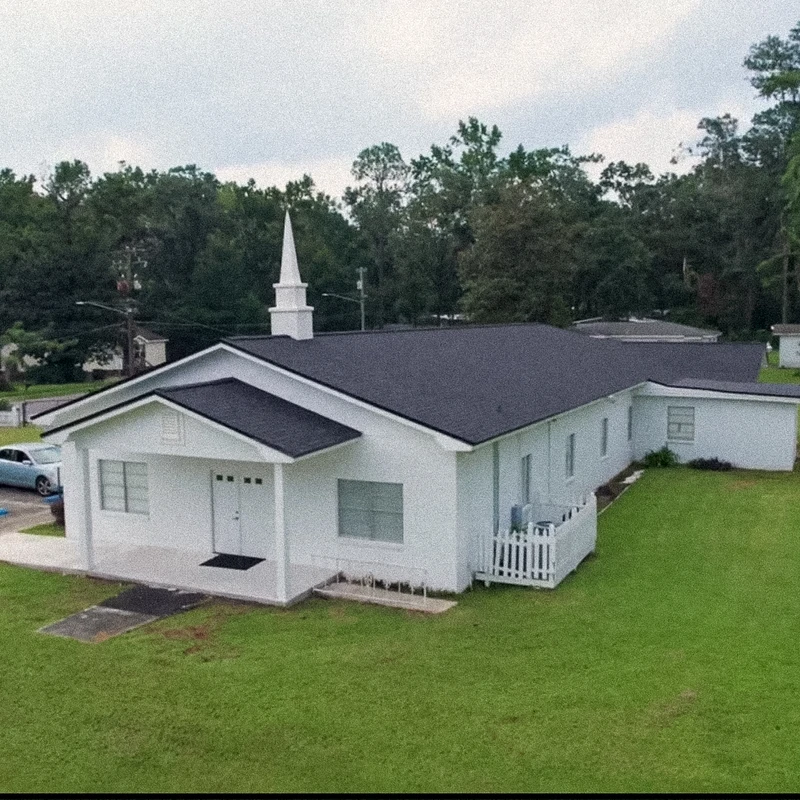 An aerial view of a single-story white church with a black roof and a steeple, set on a large green lawn with trees and houses in the background.