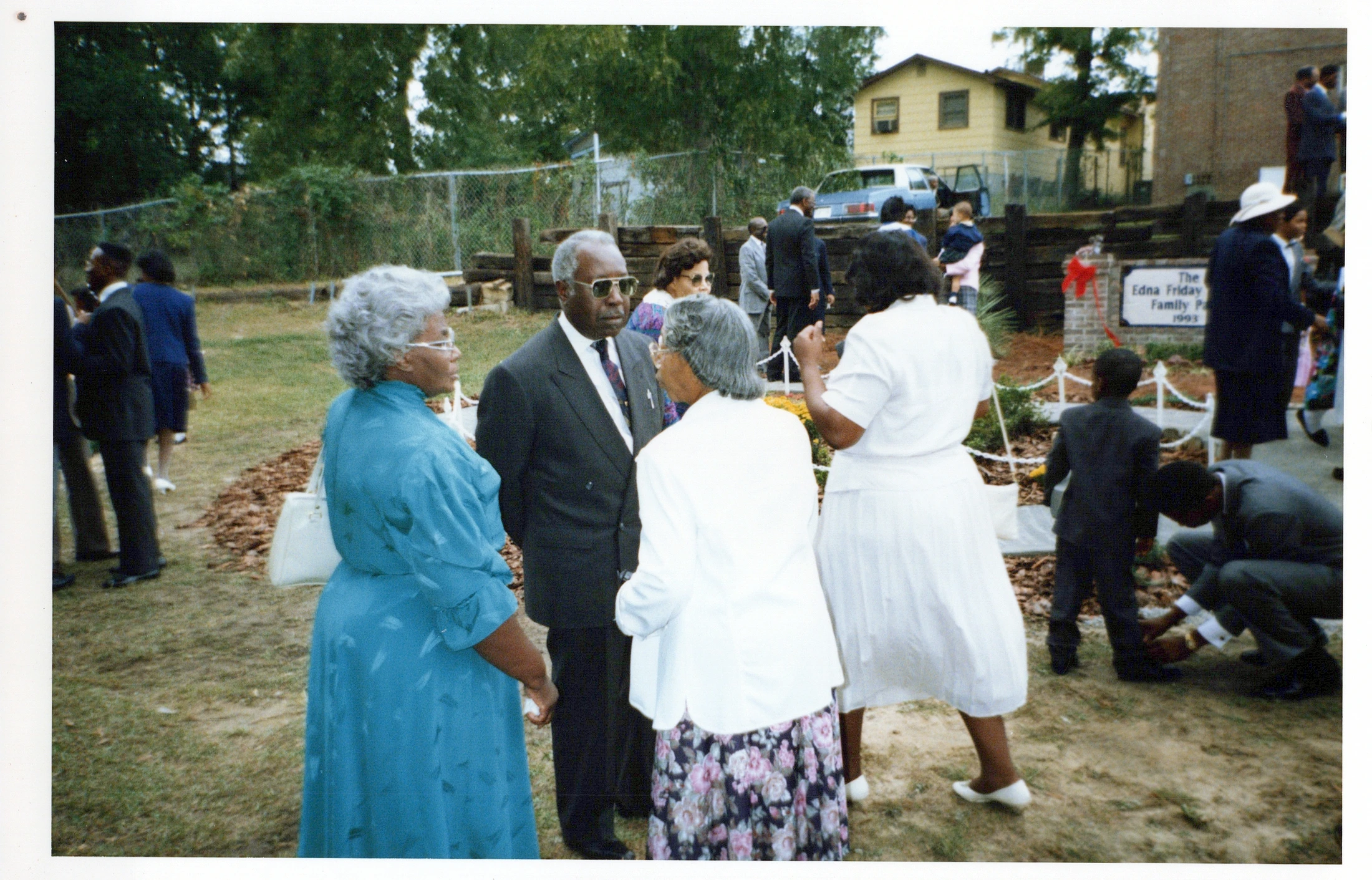 A group of well-dressed African American family members gather outdoors in 1993. Four older relatives chat in the foreground while others mingle in the back.
