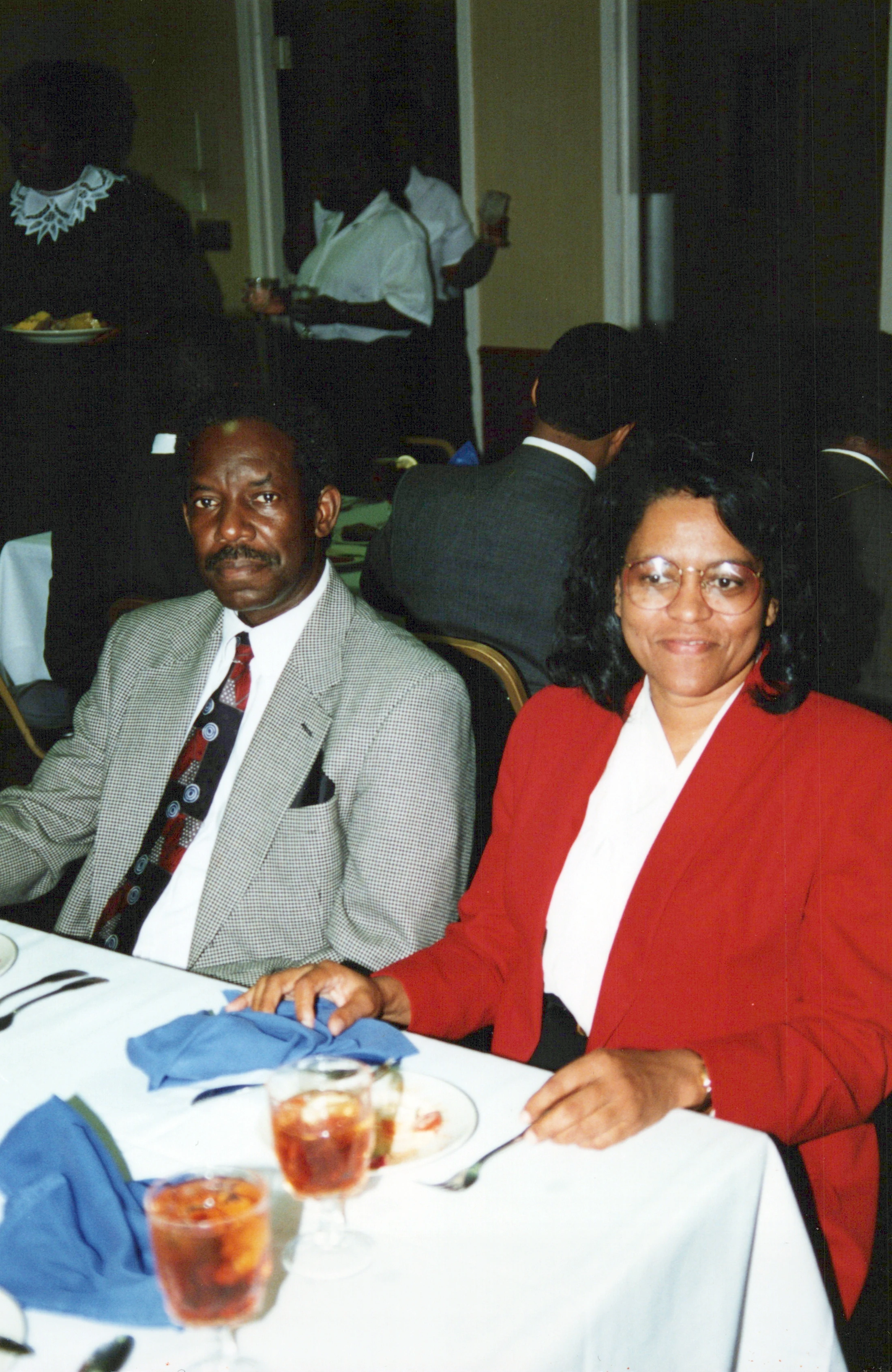 A Black man in a grey patterned jacket and a Black woman in a red blazer sit side-by-side at a formally set dinner table during an event.