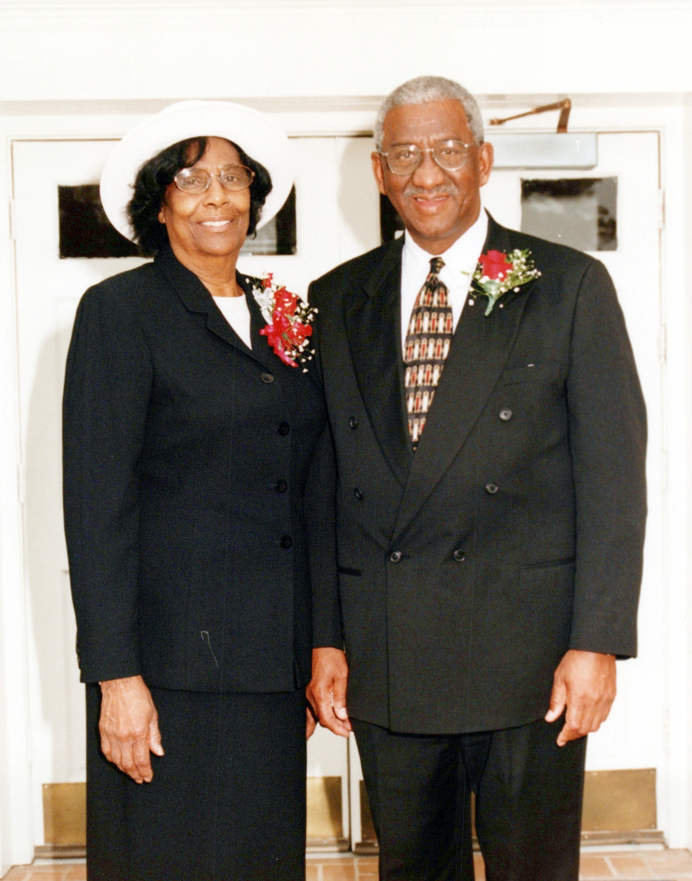 An elderly Black man and woman stand side-by-side, smiling. She wears a black suit and a white hat. He wears a black double-breasted suit and glasses.