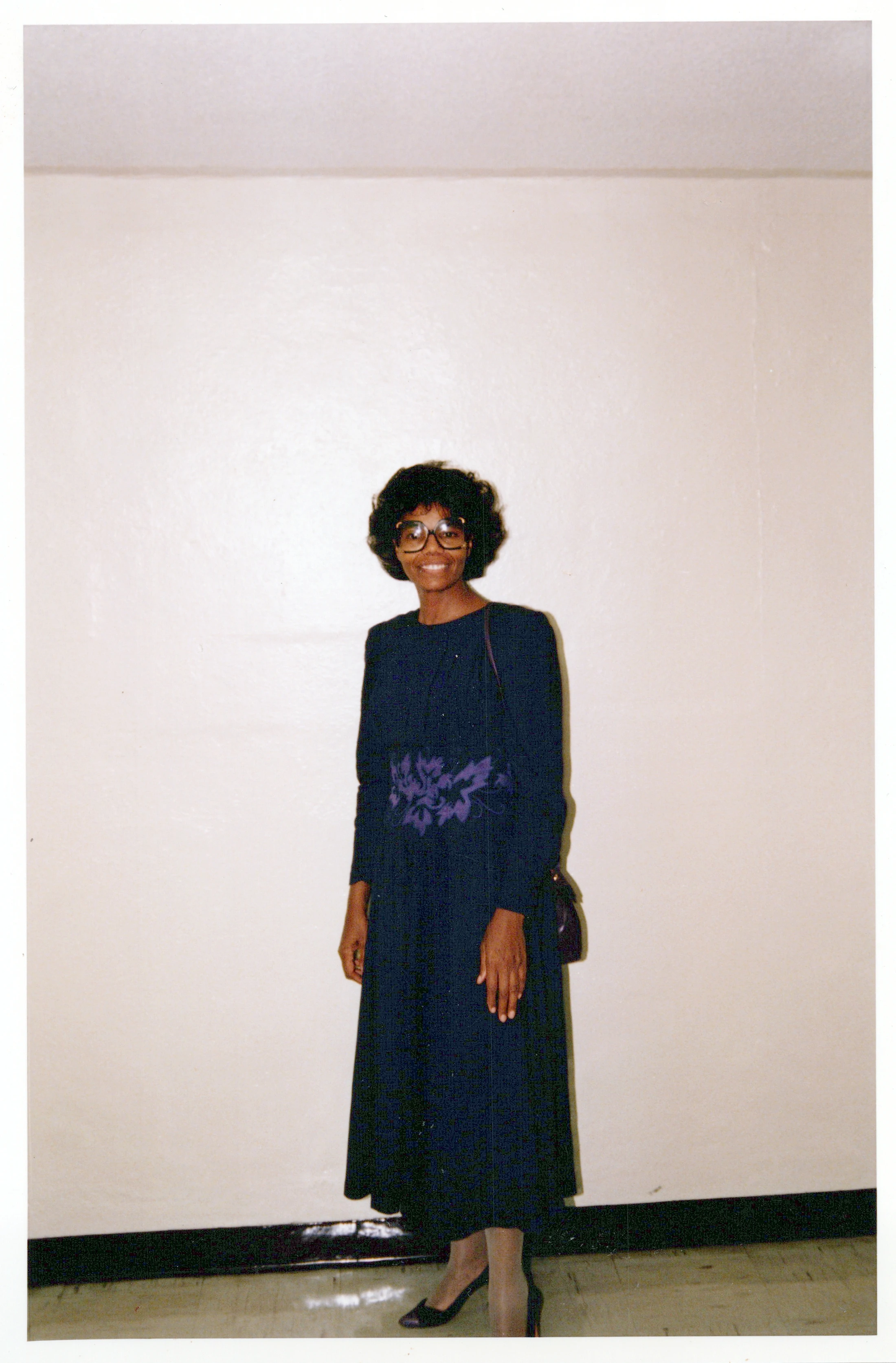 A full-length vintage photo of a Black woman with short curly hair and large glasses. She smiles while wearing a dark blue dress with a purple floral design.